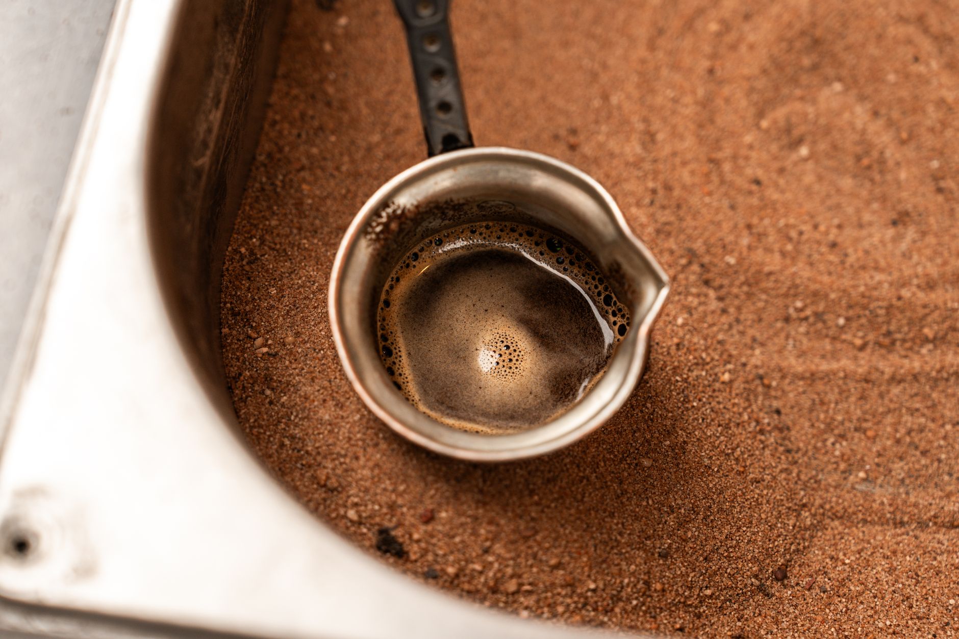 Close-up of a cezve with Turkish coffee bubbling in hot sand for brewing.