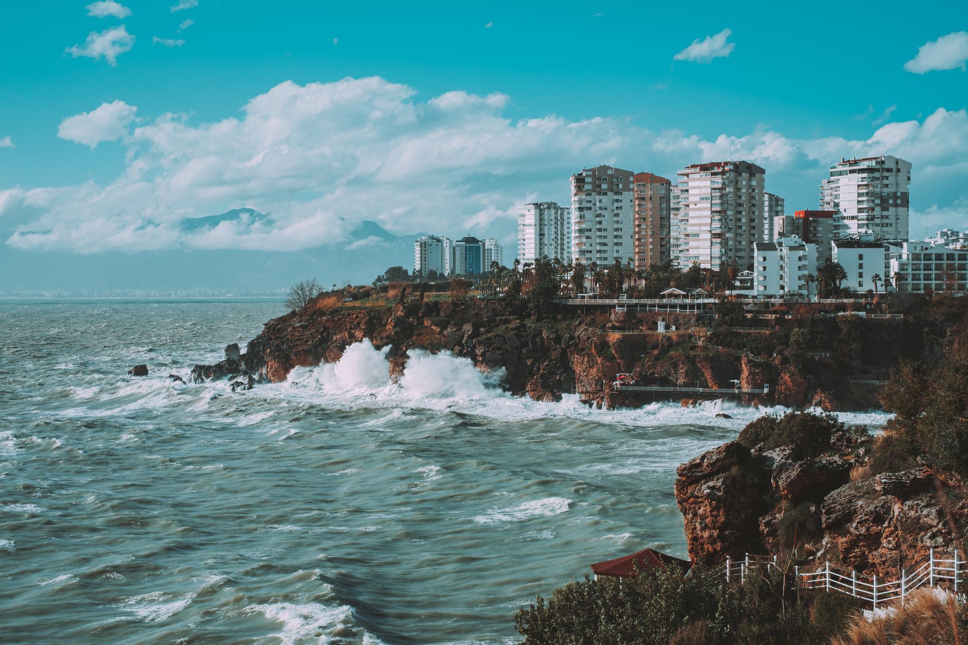 Waves crashing against the cliffs of Antalya, with modern buildings in the background and a vibrant blue sky.