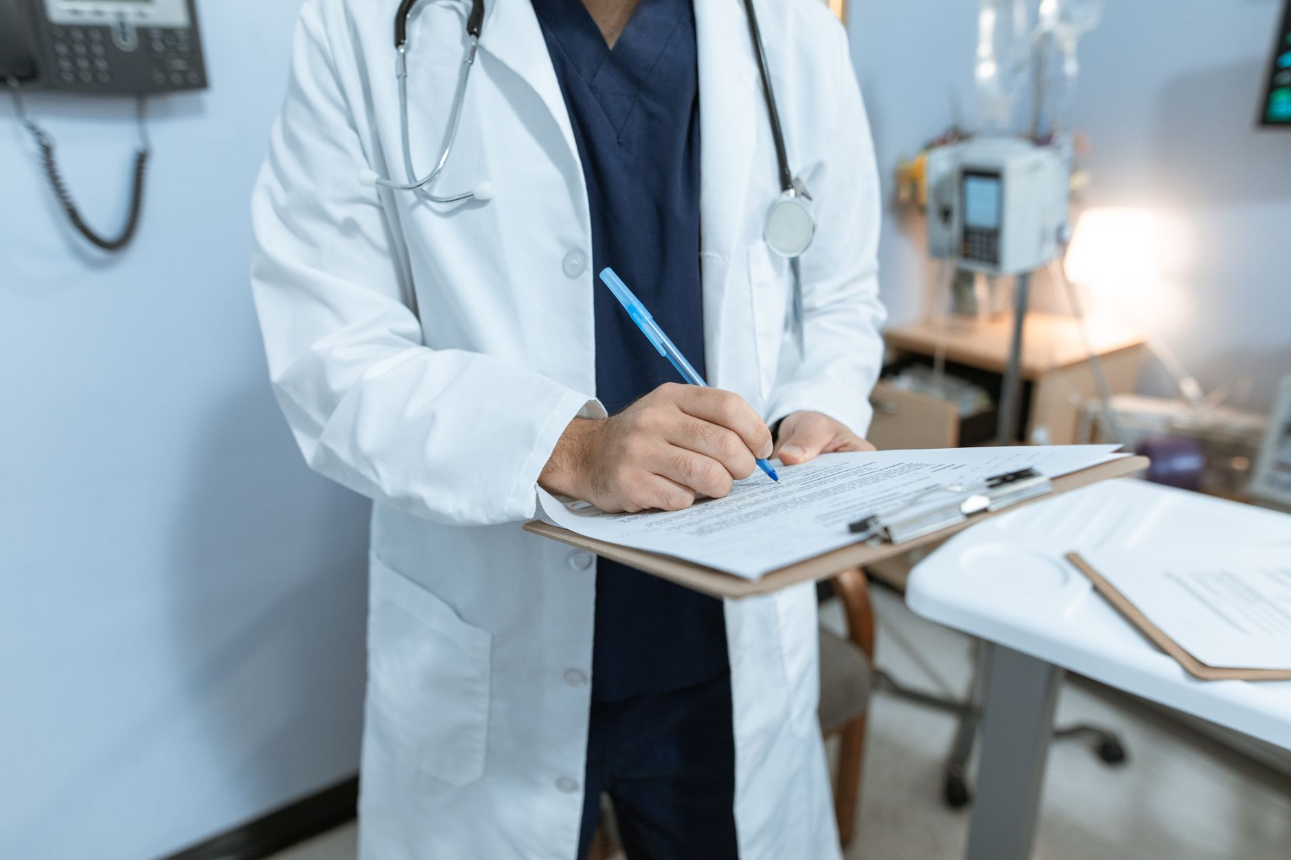 A doctor in a white coat writing on a clipboard in a hospital room.
