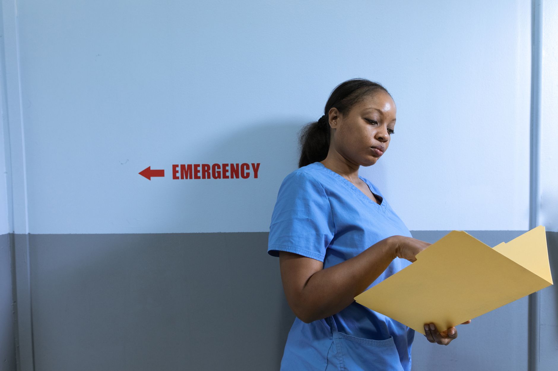 A healthcare worker in blue scrubs examines a yellow folder with medical records in a hospital corridor.