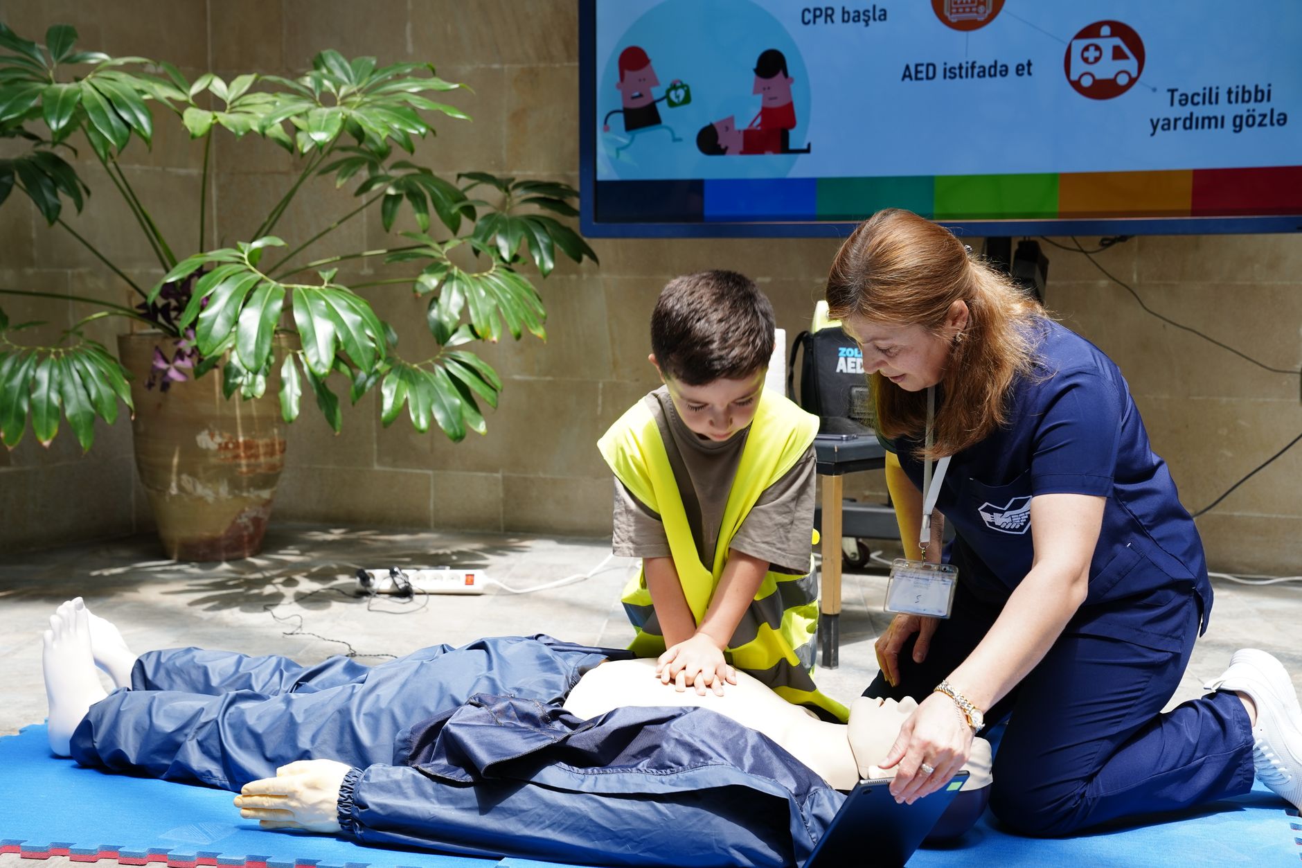 A child practices CPR on a mannequin with guidance in a hospital classroom.
