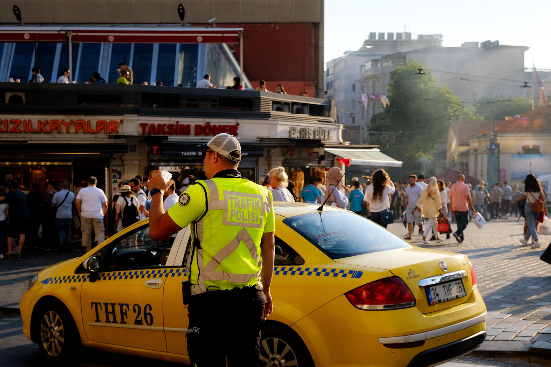 A traffic police officer directing traffic near a busy Taksim Square in Istanbul, with pedestrians and a yellow taxi.