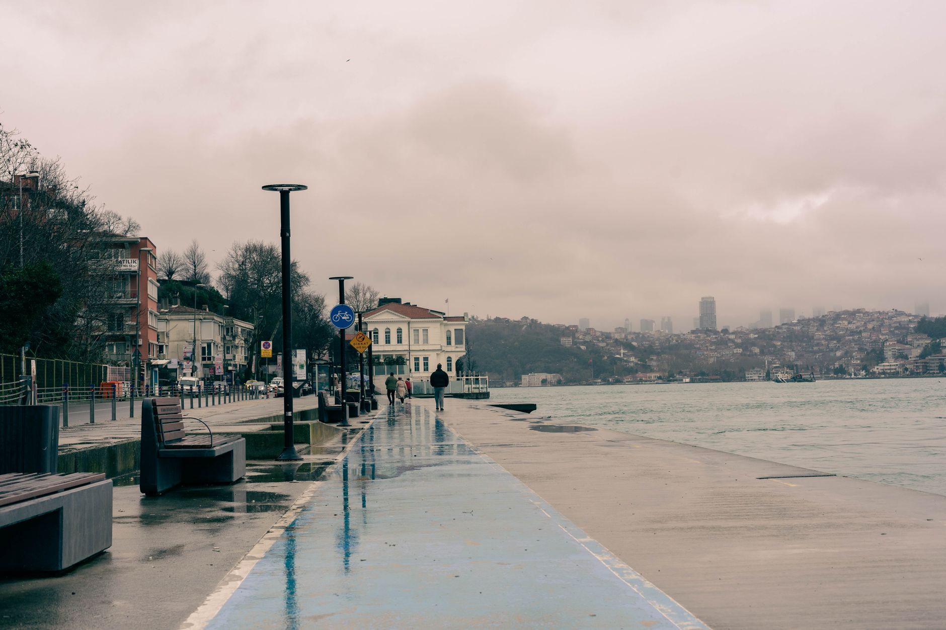 A serene Bosphorus waterfront scene in Istanbul with wet pavement and people walking.