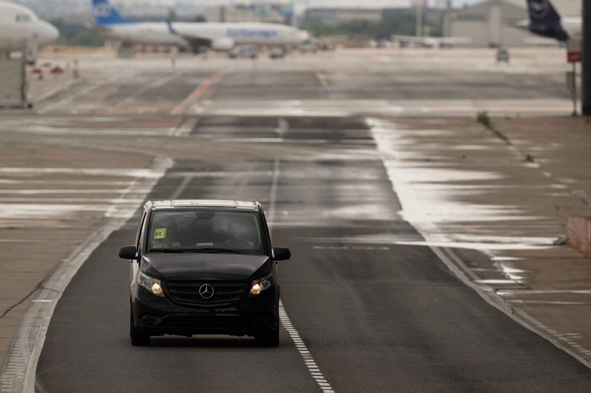 A black Mercedes vehicle driving on an airport runway with planes in the background.