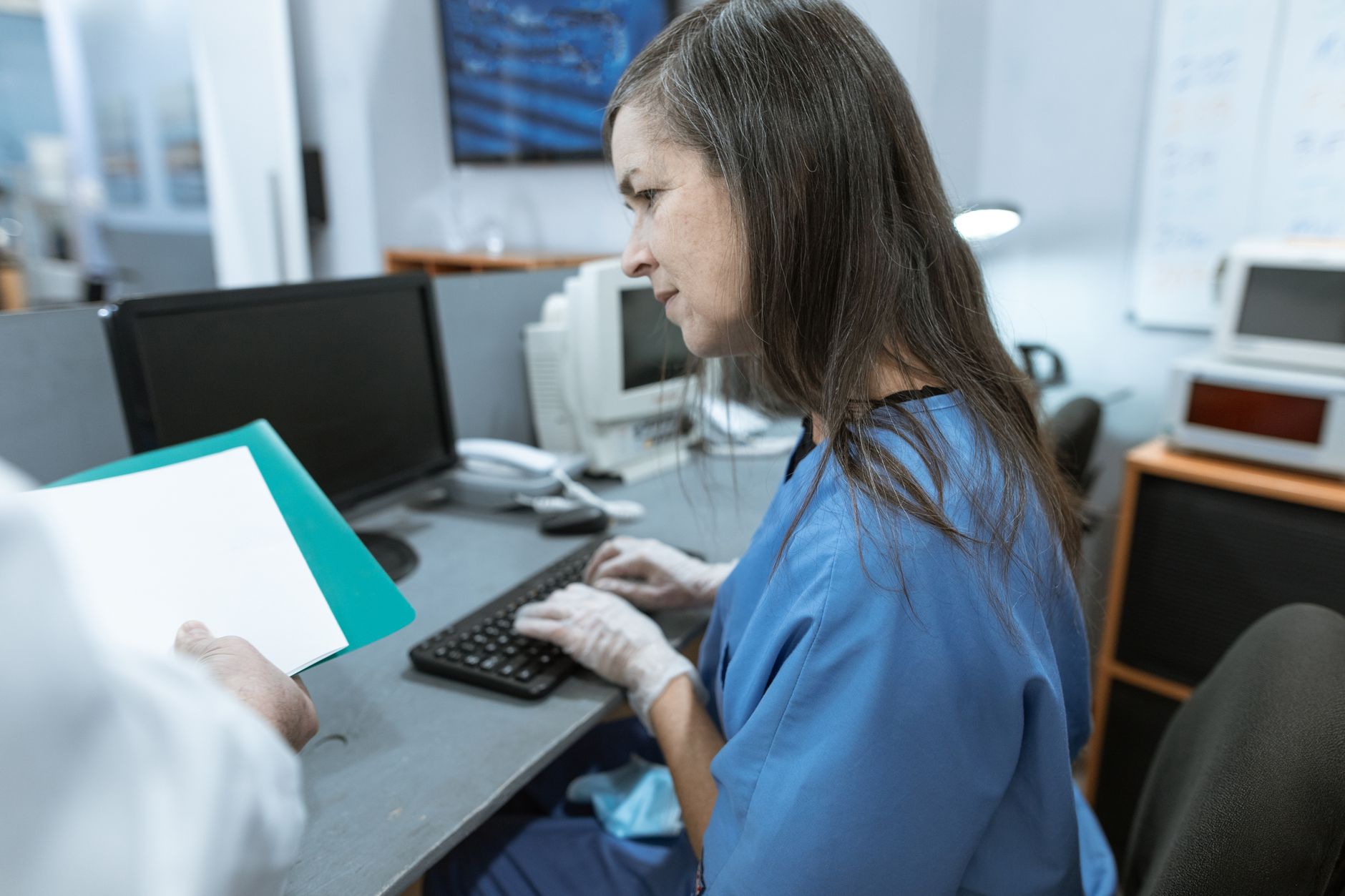 A healthcare worker in blue scrubs using a computer to manage medical records in an office environment.