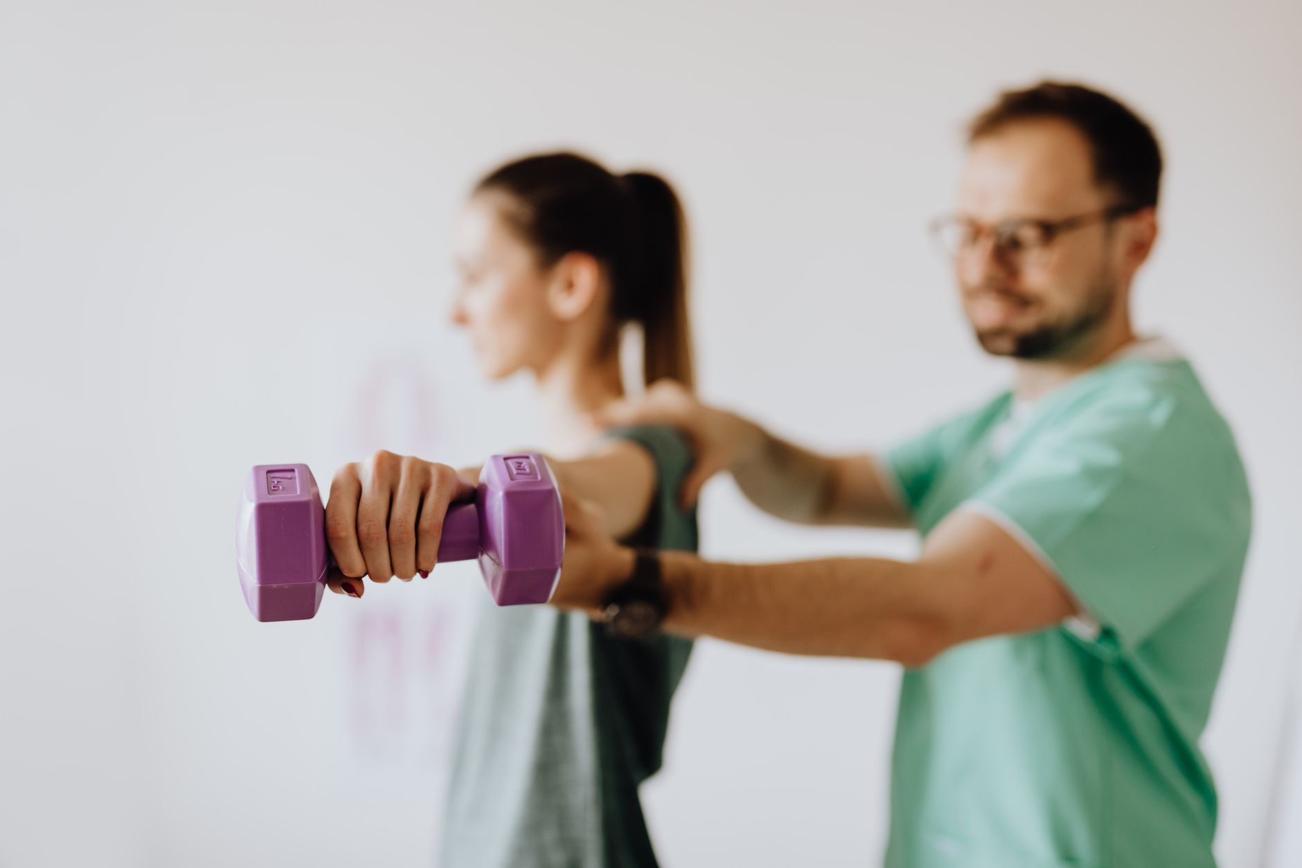 A physiotherapist guides a patient using a purple dumbbell during rehabilitation in a bright clinic.