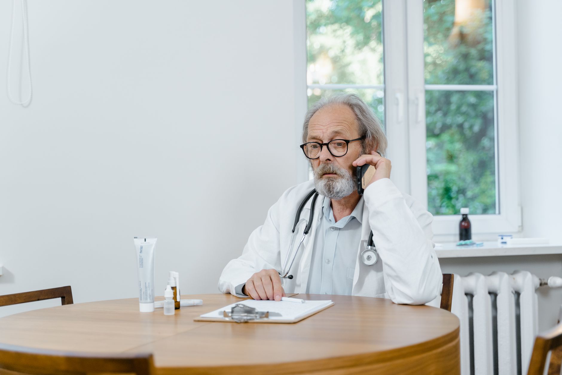 An older doctor in a white coat speaking on the phone at a wooden table with medical supplies in an office.