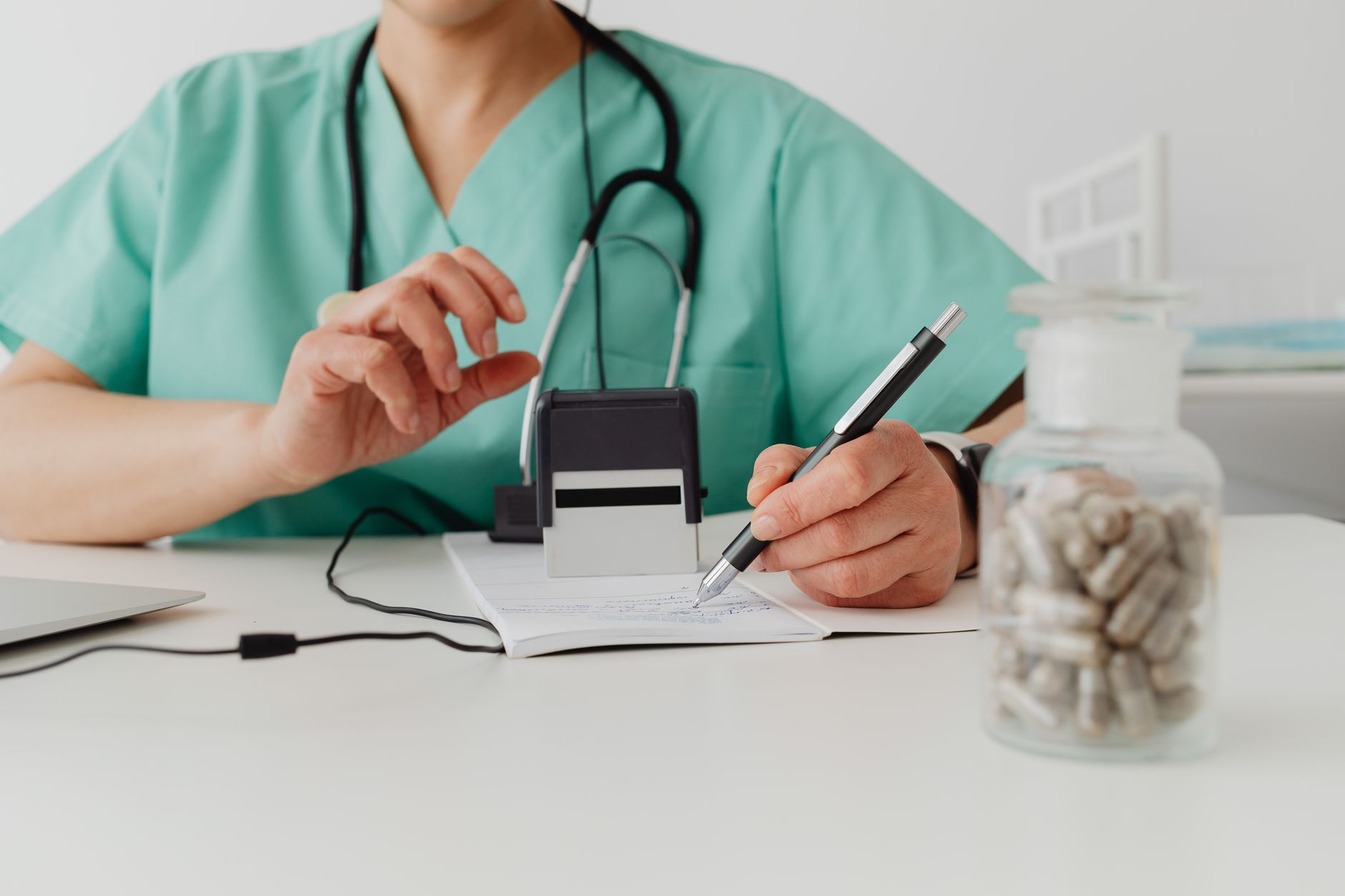 A doctor in scrubs writing notes with a stethoscope and a bottle of pills nearby.