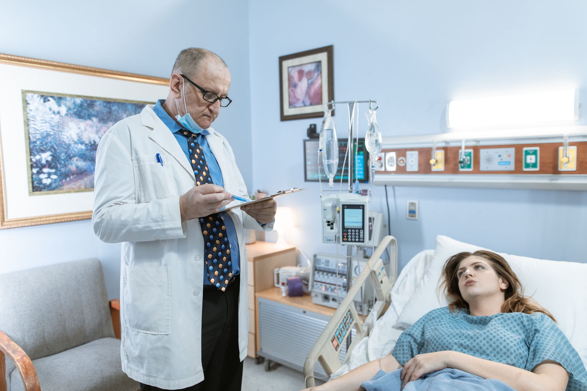 A doctor in a white coat reviews a clipboard beside a recovering patient in a hospital room.