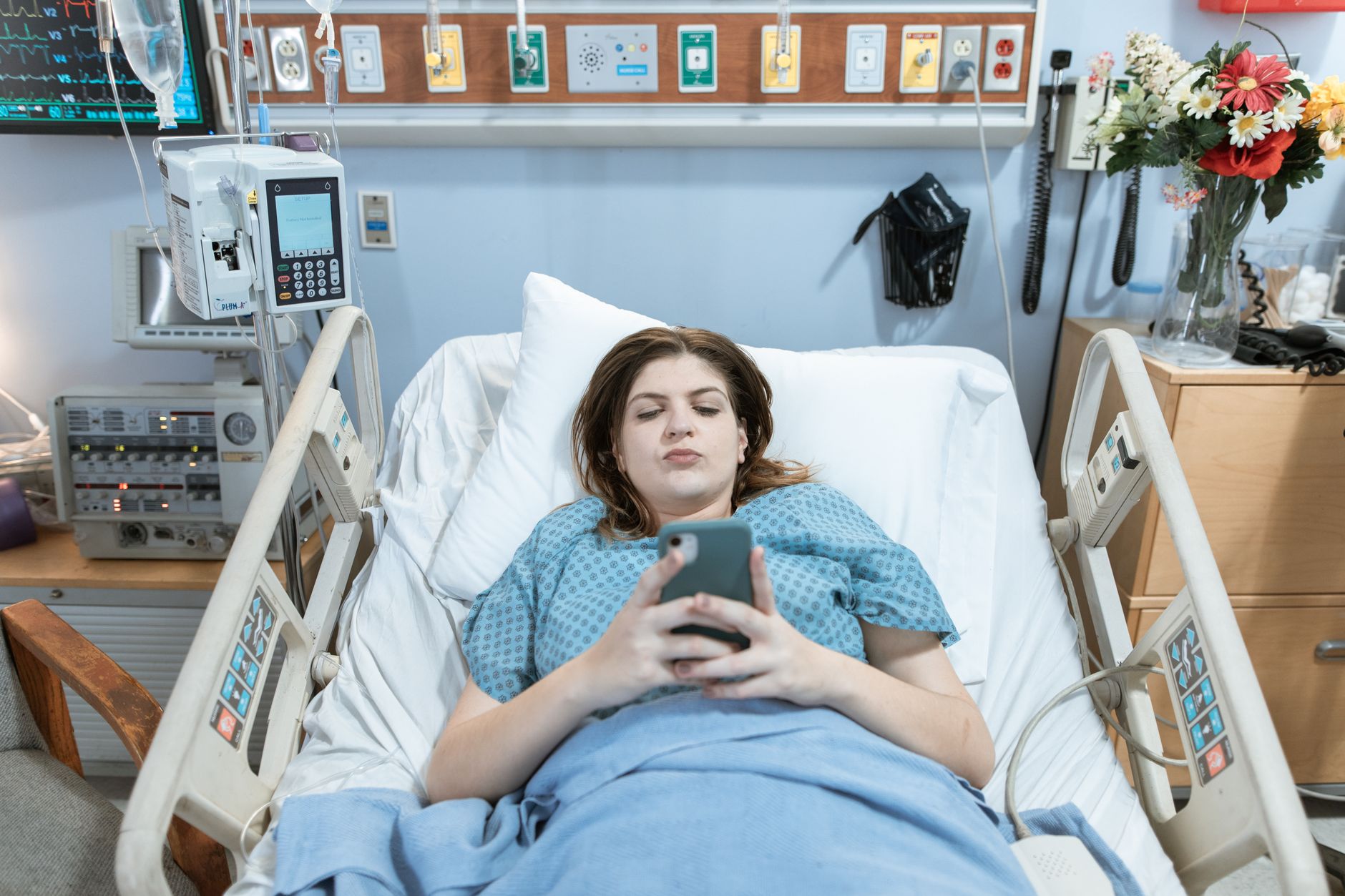 A patient in a hospital bed using a smartphone while connected to an infusion pump.