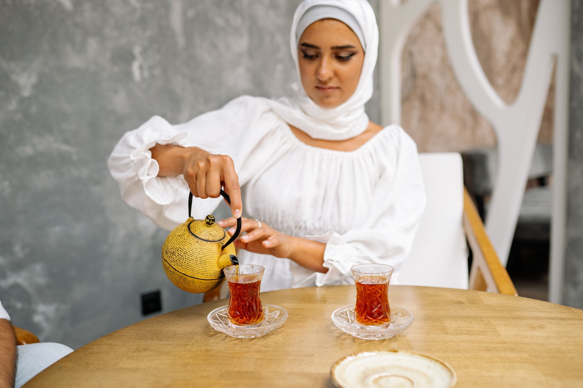 زنی با لباس سفید چای ترکی را از قوری زردی در استکان‌های شیشه‌ای می‌ریزد.