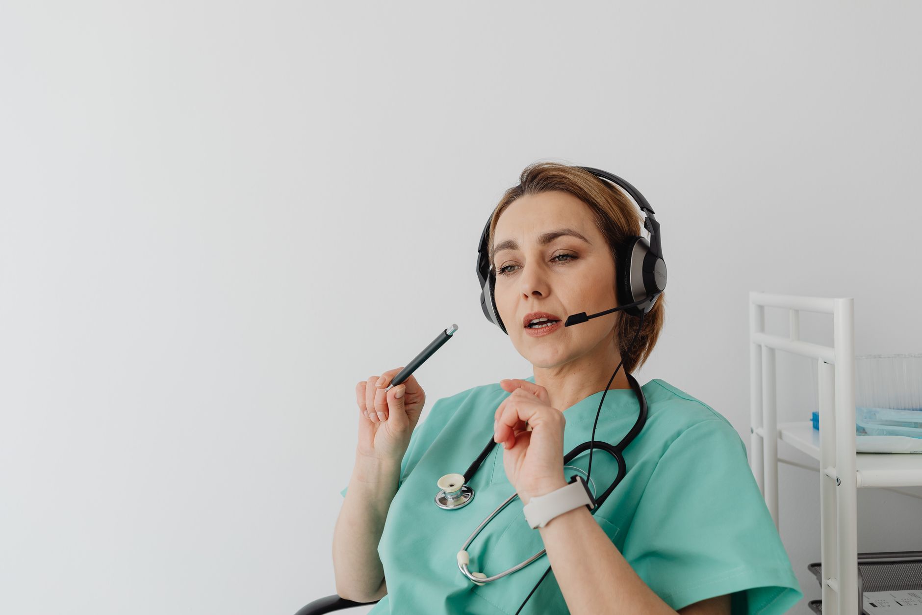 Healthcare provider in scrubs using a headset for a telemedicine consultation, holding a pen.