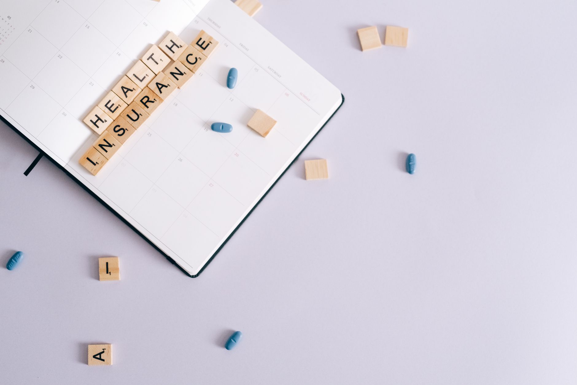 Wooden tiles spelling 'Health Insurance' over a blank calendar with blue pills.
