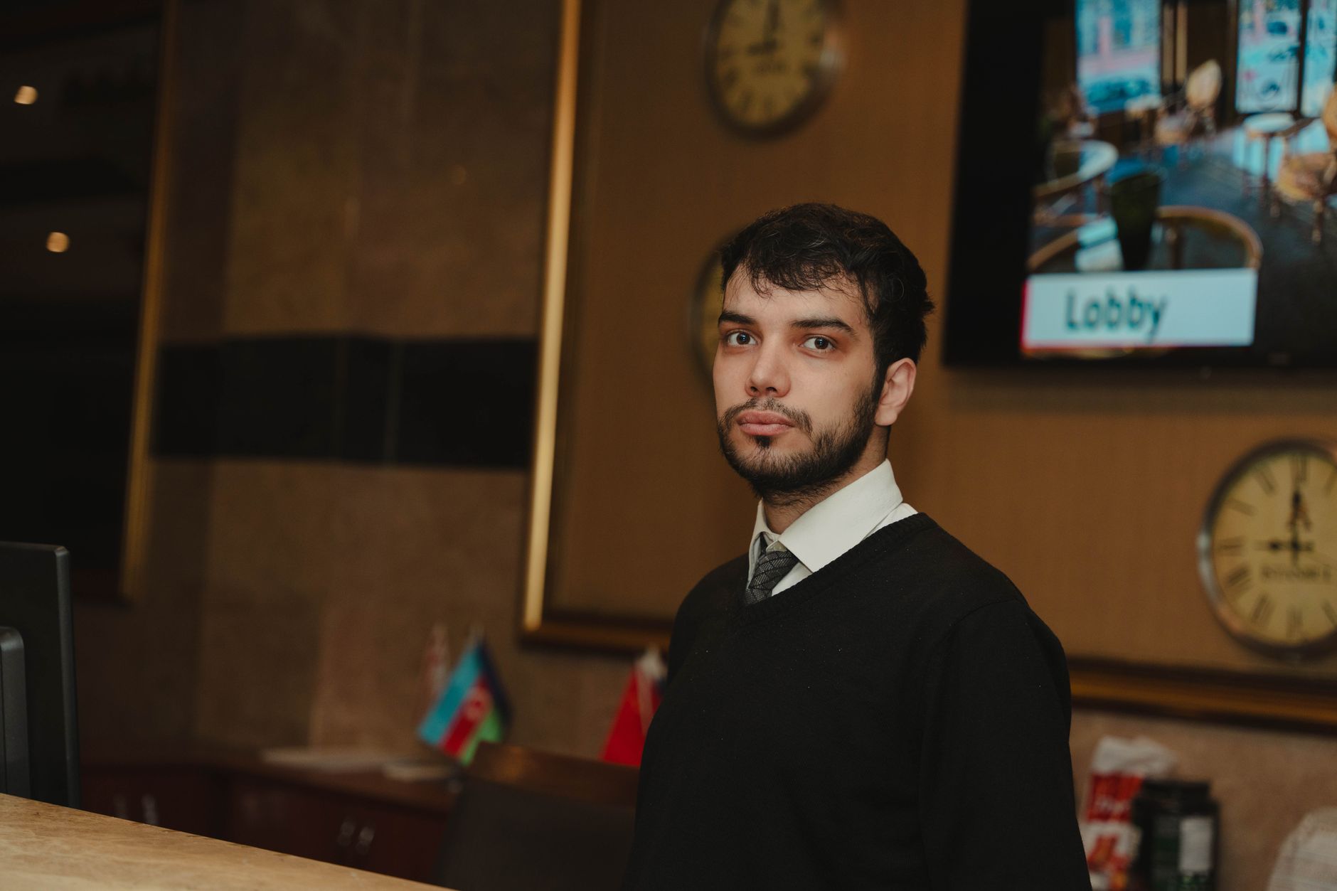 A hotel receptionist in a black sweater aiding guests. Flags of Azerbaijan and Georgia visible in the background.