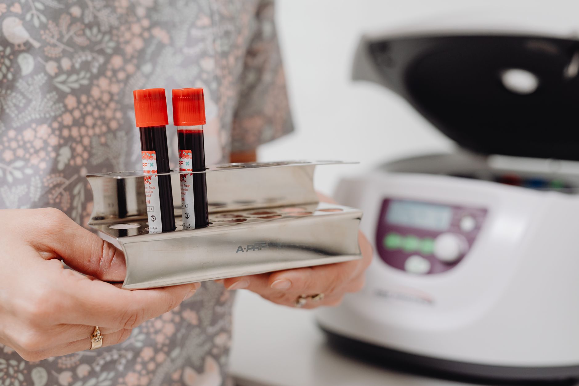 Nurse holding blood sample tubes in a stainless steel rack near a centrifuge.