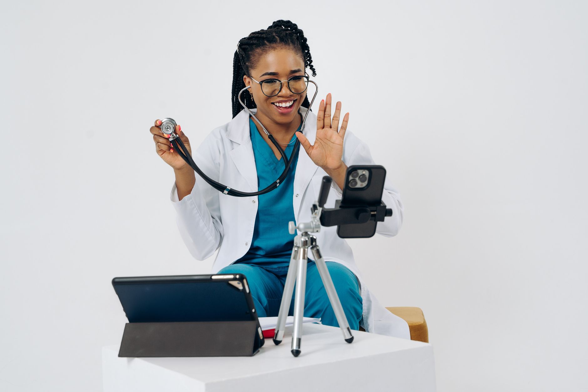 Doctor in white coat waves while using stethoscope in a telehealth setup.