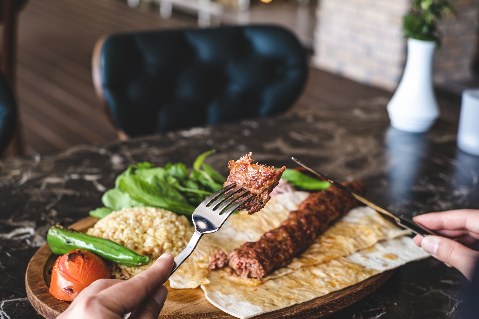 A fork holding a piece of kebab alongside rice, greens, and grilled vegetables on a wooden platter.