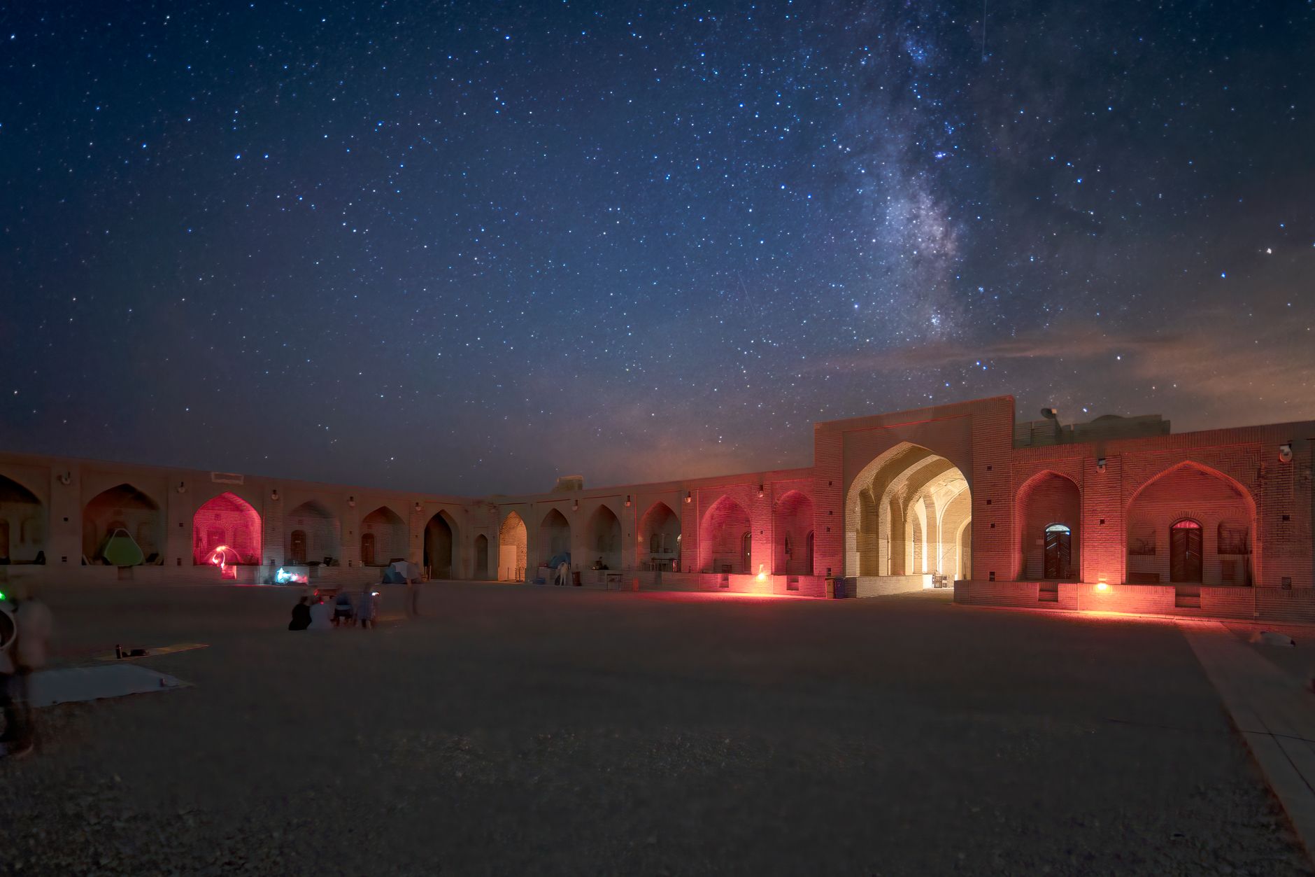 نمای شبانه حیاط یک کاروانسرای بازسازی‌شده با رواق‌ها و مسجدی زیر آسمان پرستاره.