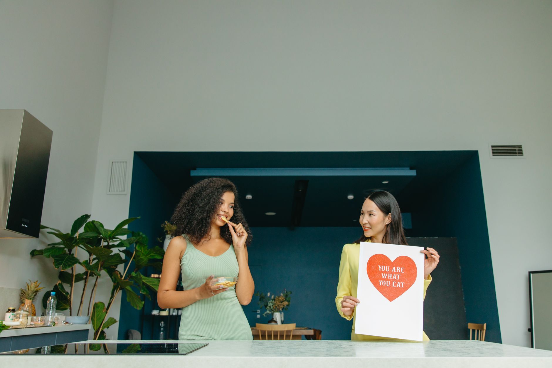 Two women in a kitchen, one enjoying a snack and the other holding a sign about healthy eating.