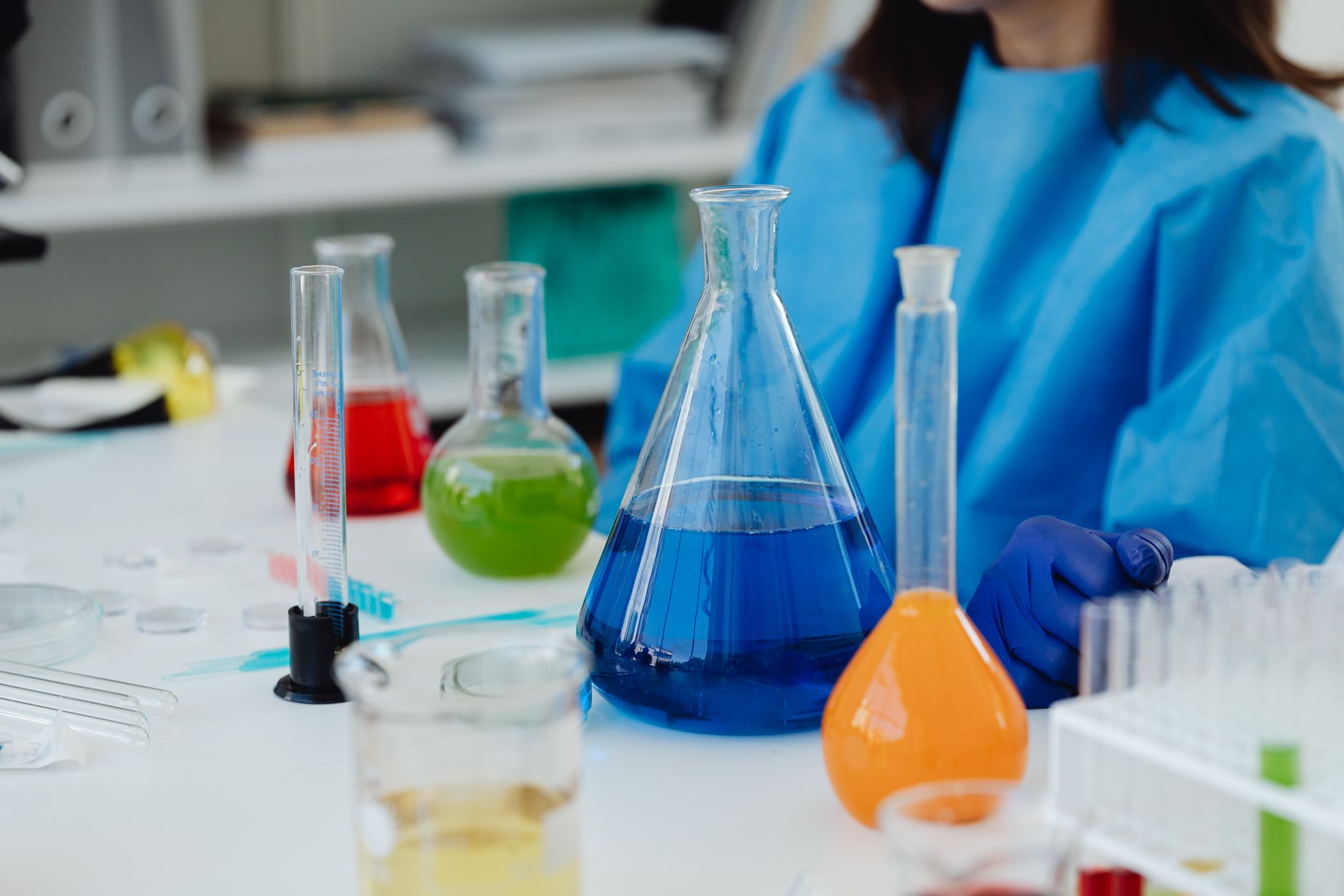 Scientist in a blue gown handling colorful flasks and beakers in a modern lab.