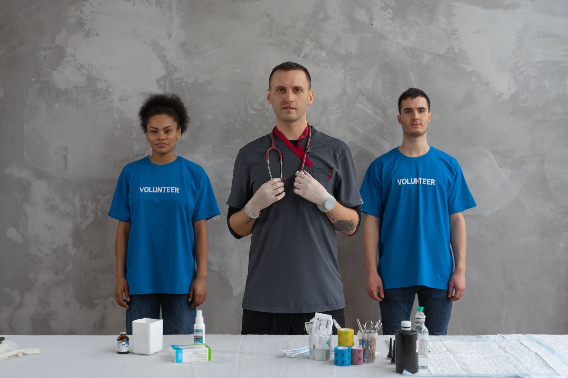 Three individuals in blue volunteer shirts assist in a healthcare setting with medical supplies.