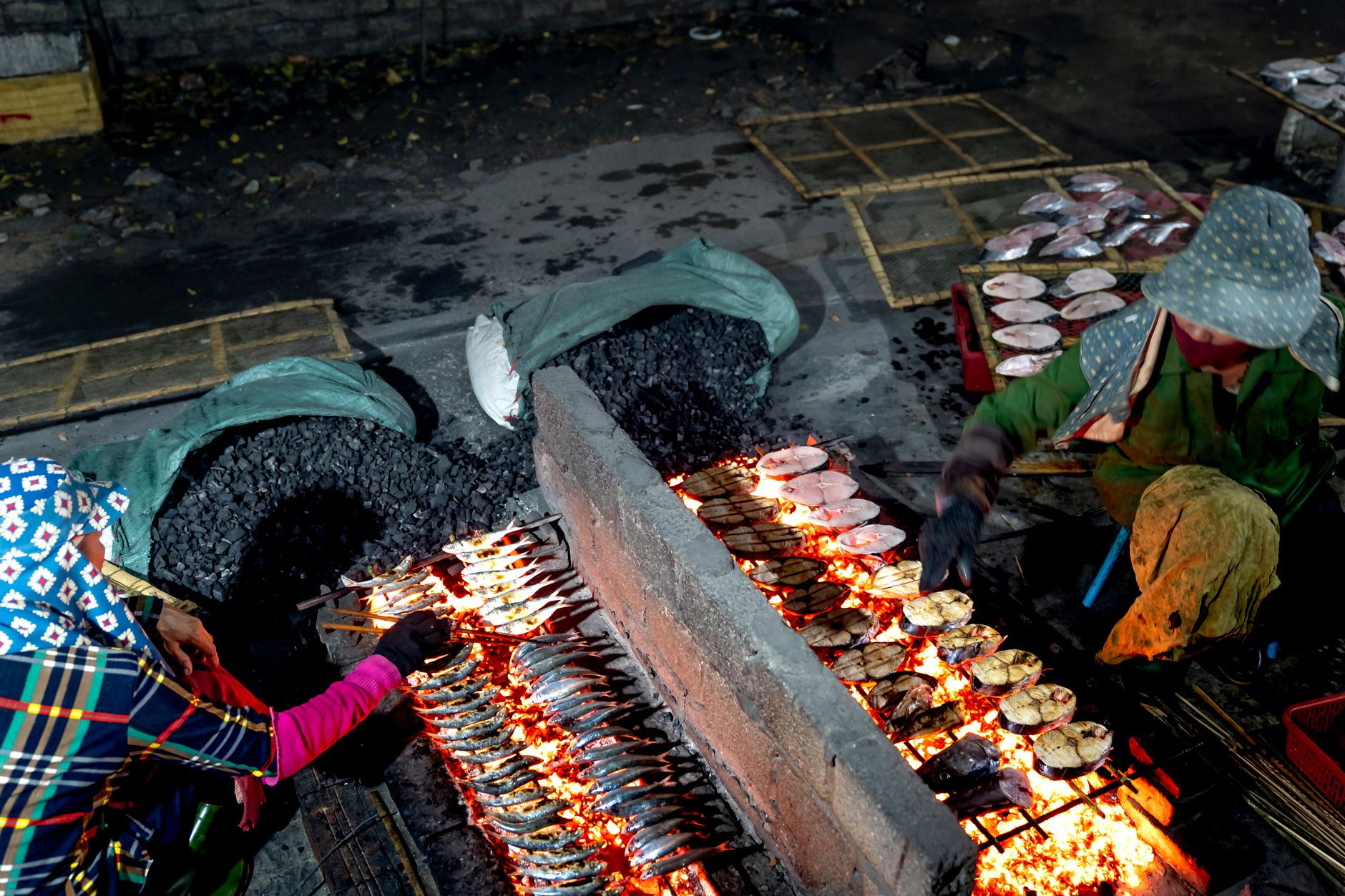 Vendors grilling fish over charcoal at a night market, with a variety of seafood visible.