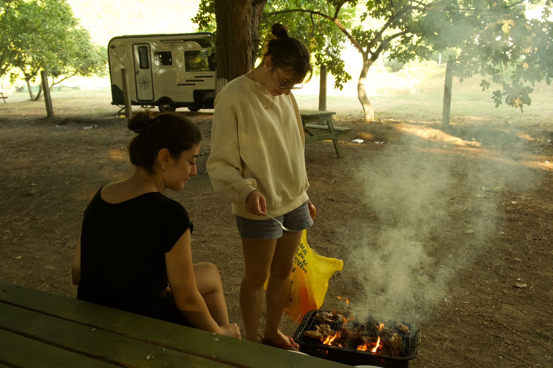 Two people cooking on a grill at a campsite near Gökova Bay, surrounded by smoke and trees.