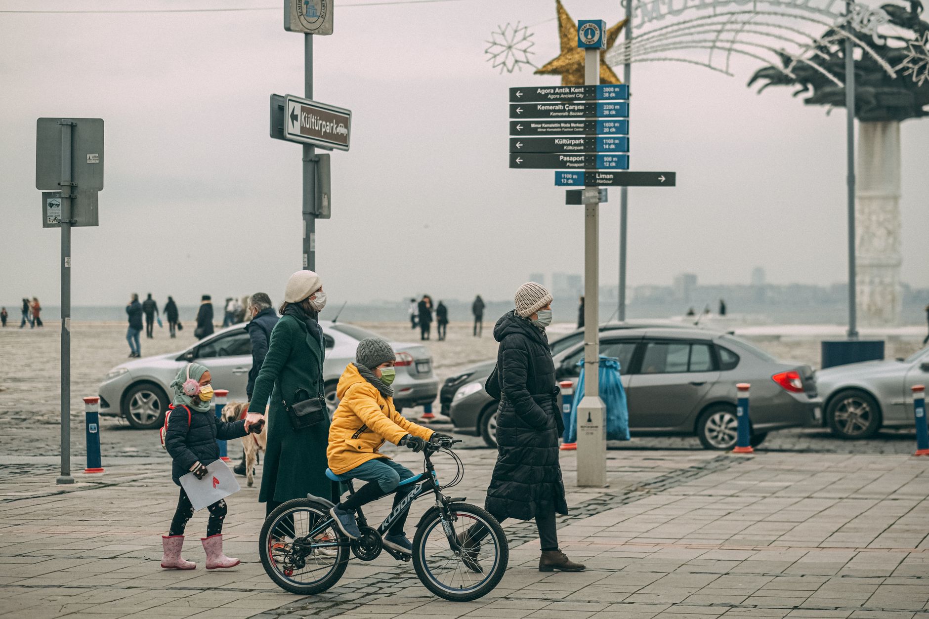A family walks along a seaside promenade with children on bicycles, wearing masks during a chilly day.