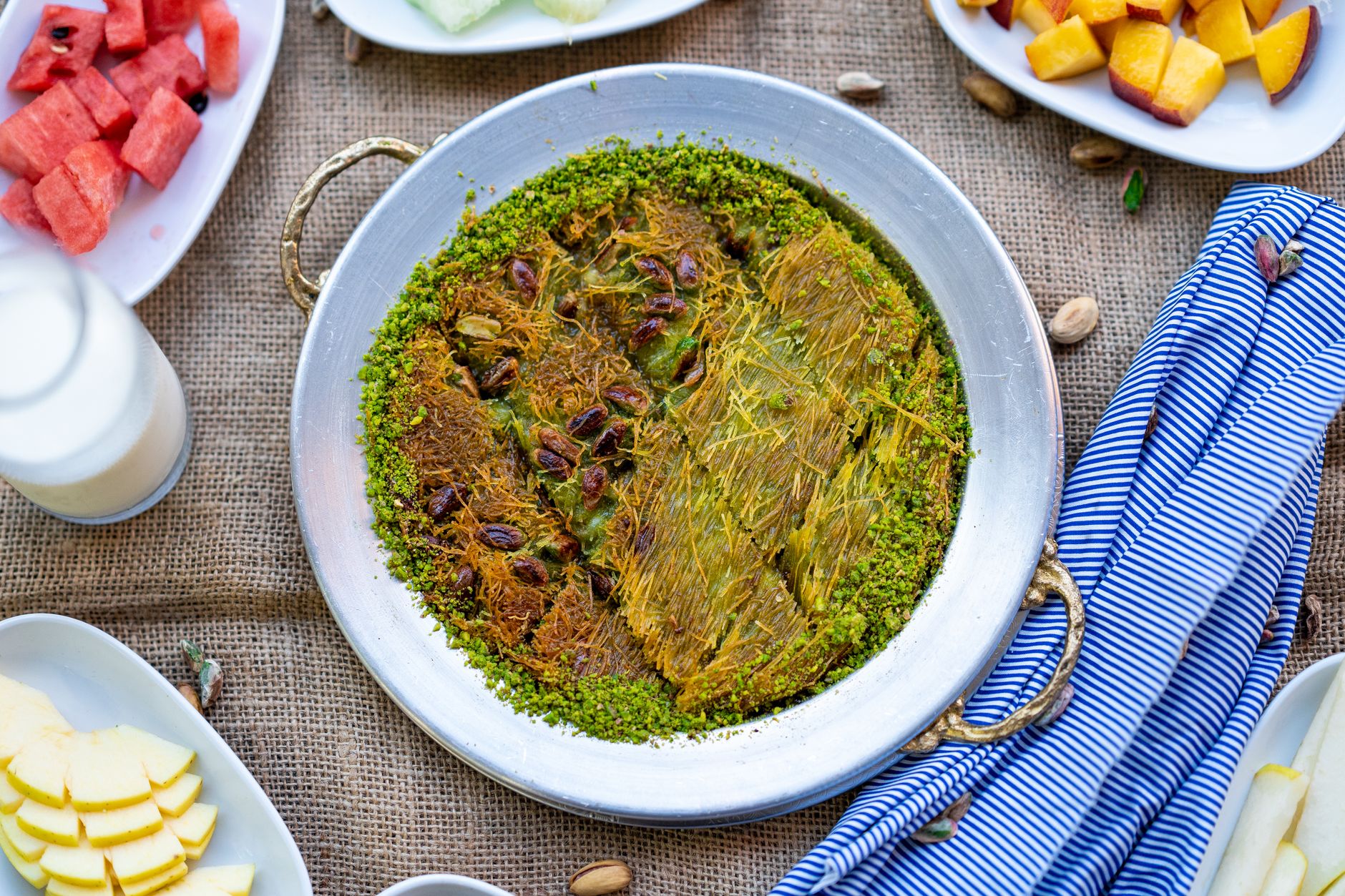 سینی مسی کونفه با شربت، پنیر و پسته، احاطه‌شده با میوه و شیر.