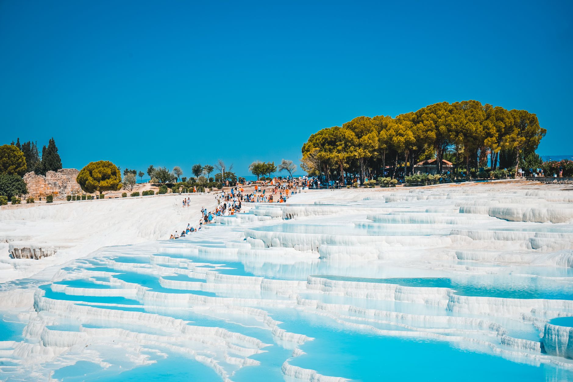 Crowds walking on the beautiful white terraces of Pamukkale, with vibrant blue water and lush greenery.