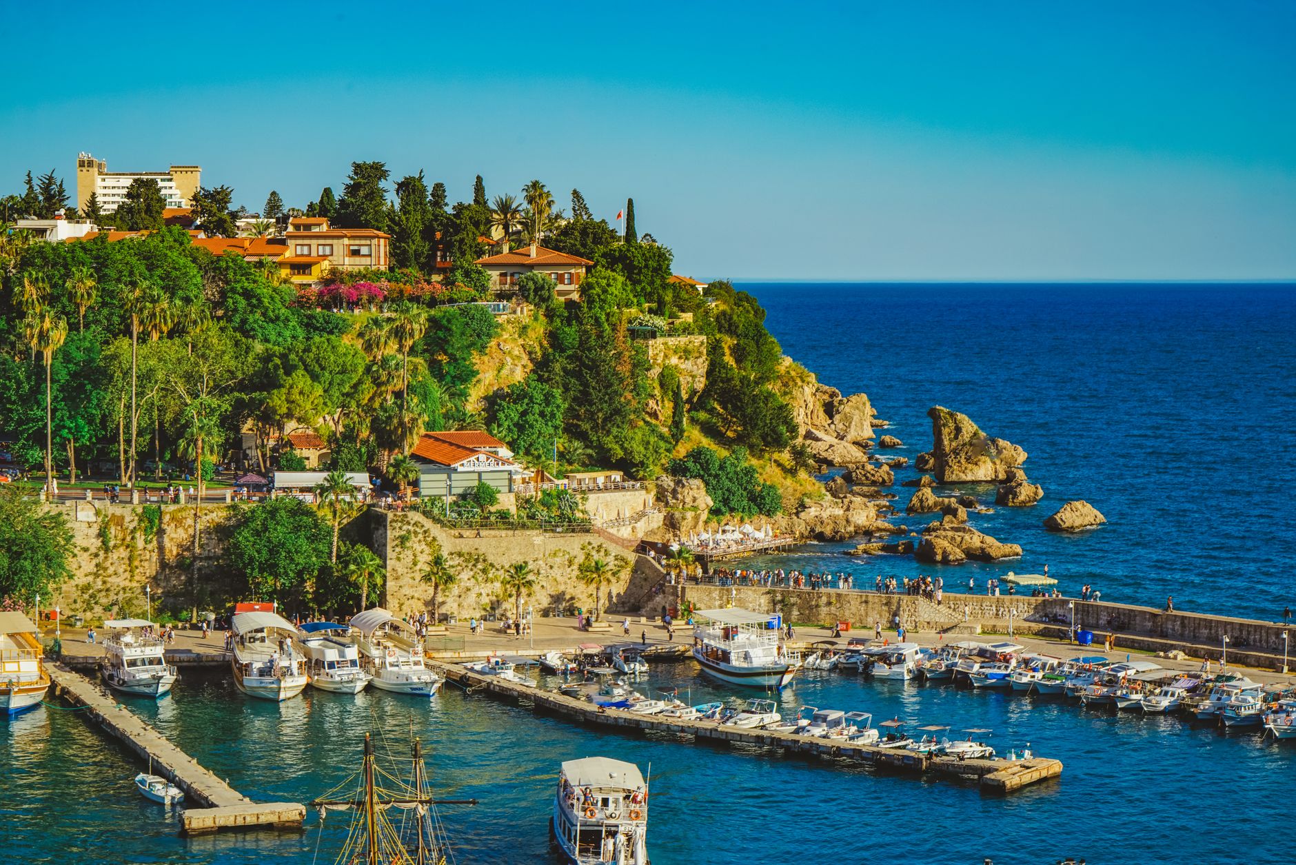A beautiful view of Antalya's harbor, showcasing boats, rocky coastline, and lush greenery.