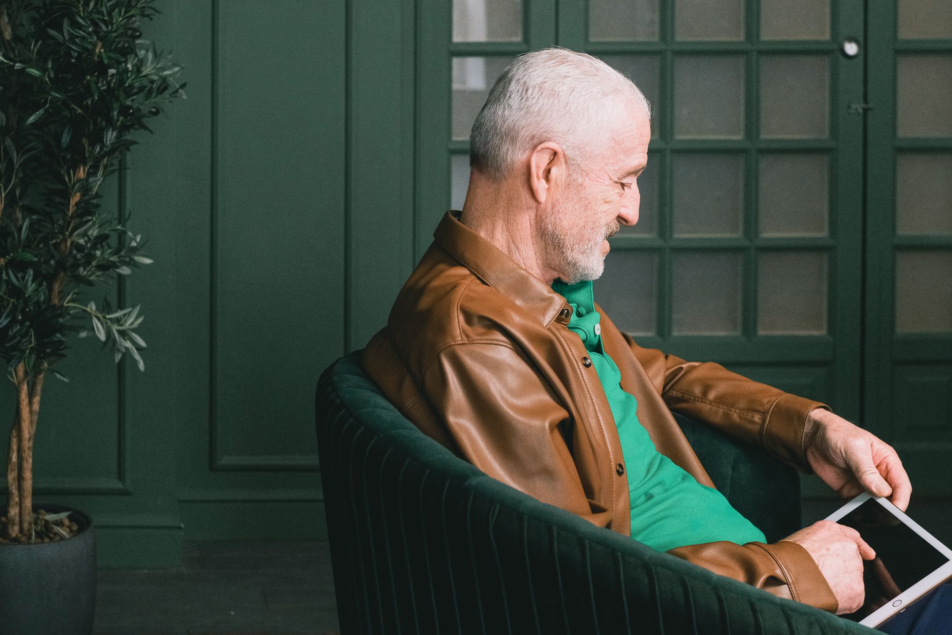 An older man in a brown leather jacket sitting in a green chair, using a tablet.