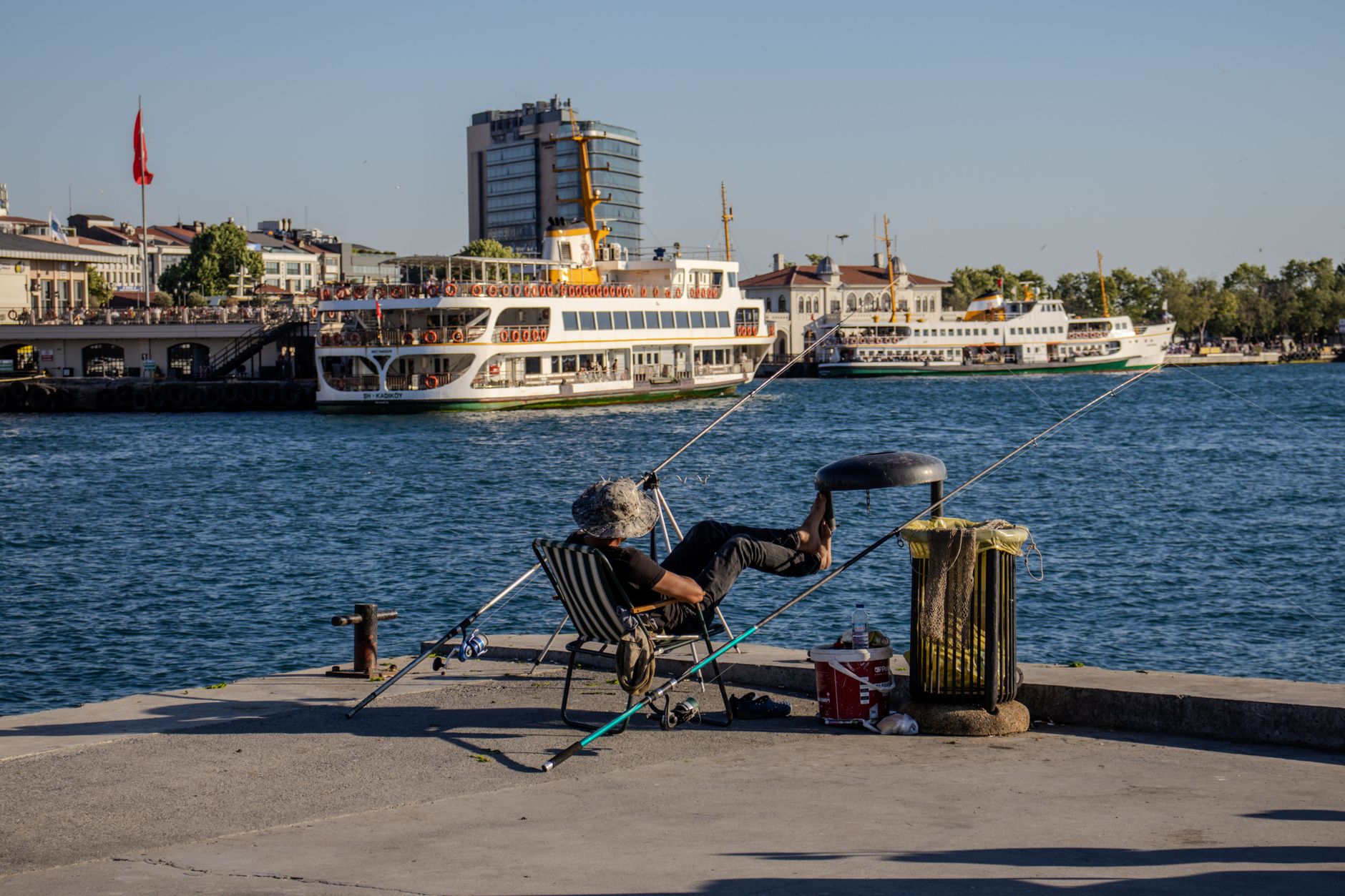 A fisherman relaxes in a chair by the Bosphorus, with ferries nearby.