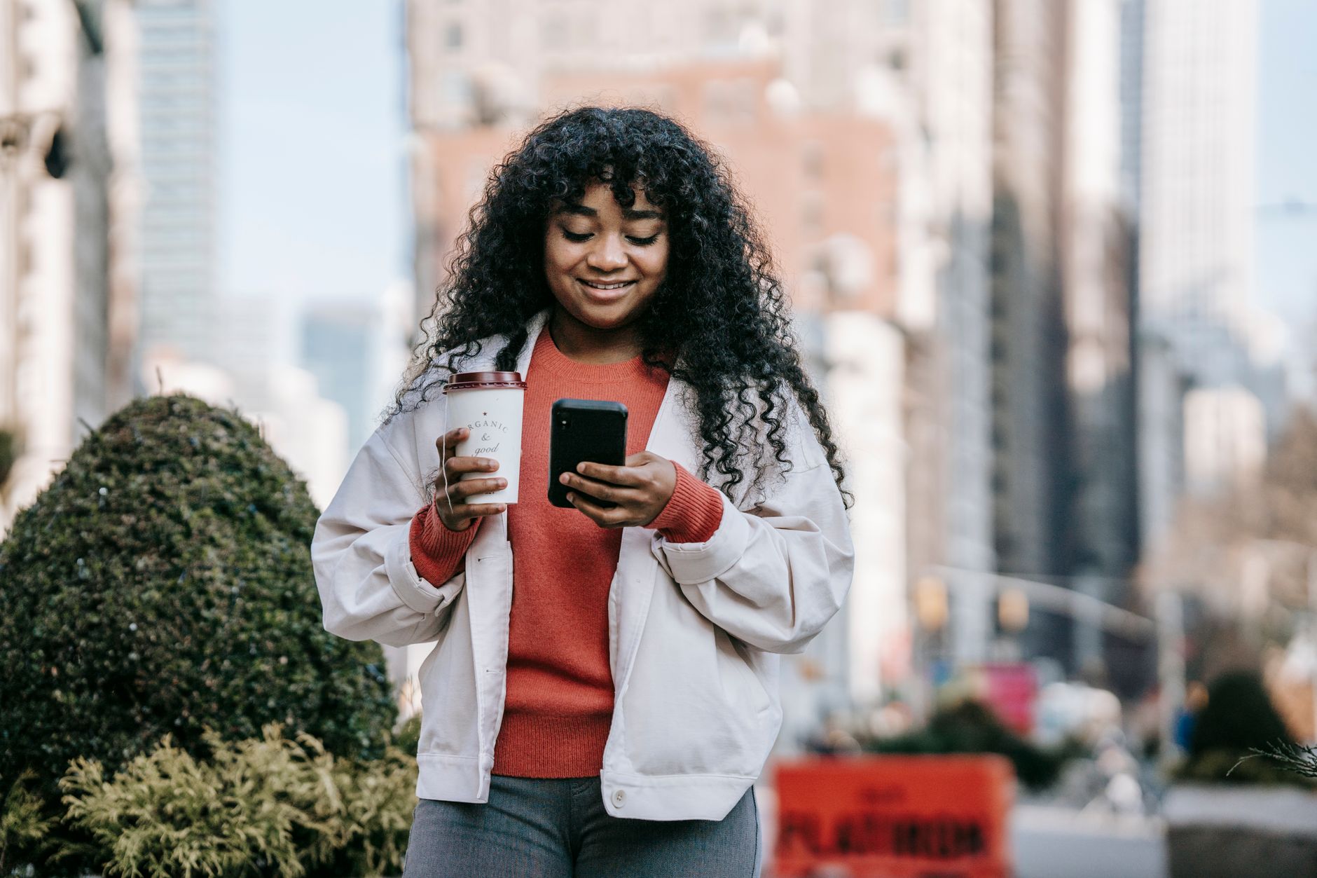 Smiling woman holding coffee and smartphone, standing outdoors in a city.
