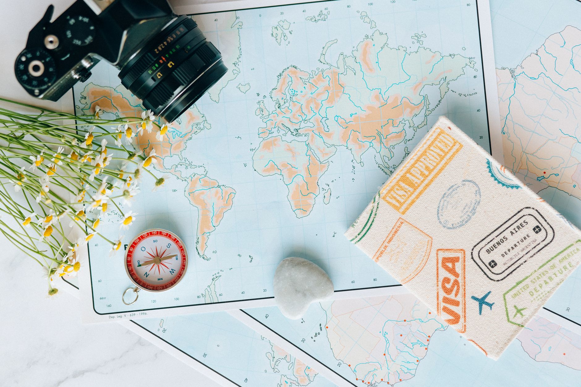 Flat lay of a world map, camera, compass, flowers, and a passport.