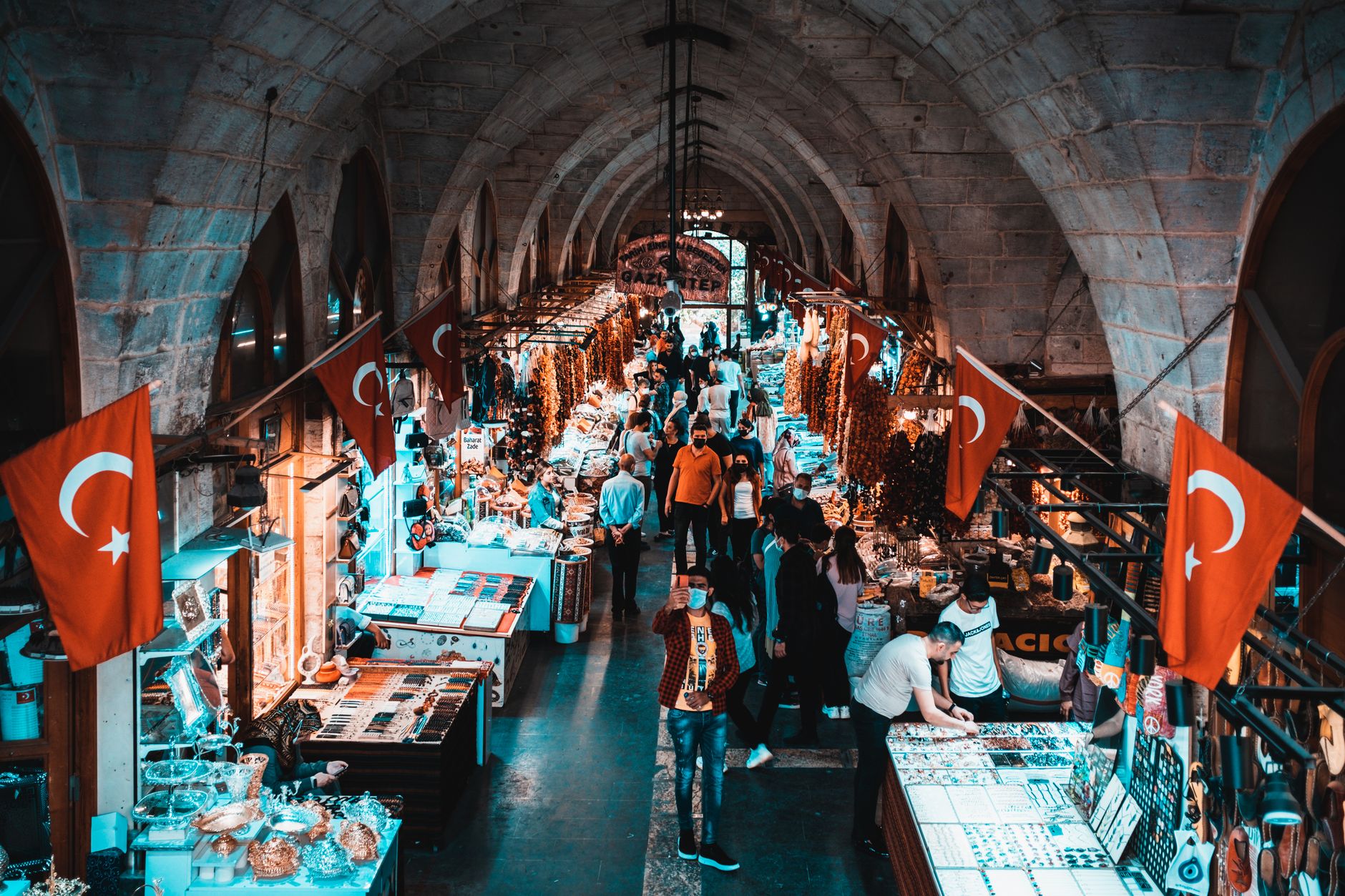 Crowd exploring colorful spice and dried vegetable stalls in Gaziantep's Elmacı Pazarı, adorned with Turkish flags.