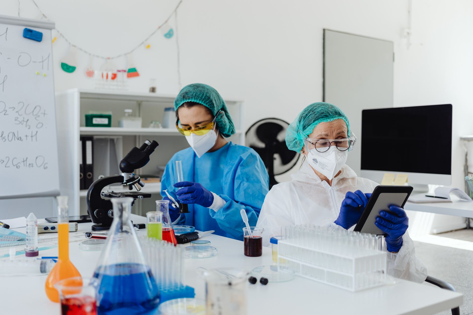 Two scientists work in a Turkish laboratory, using a microscope and tablet among colorful chemicals.