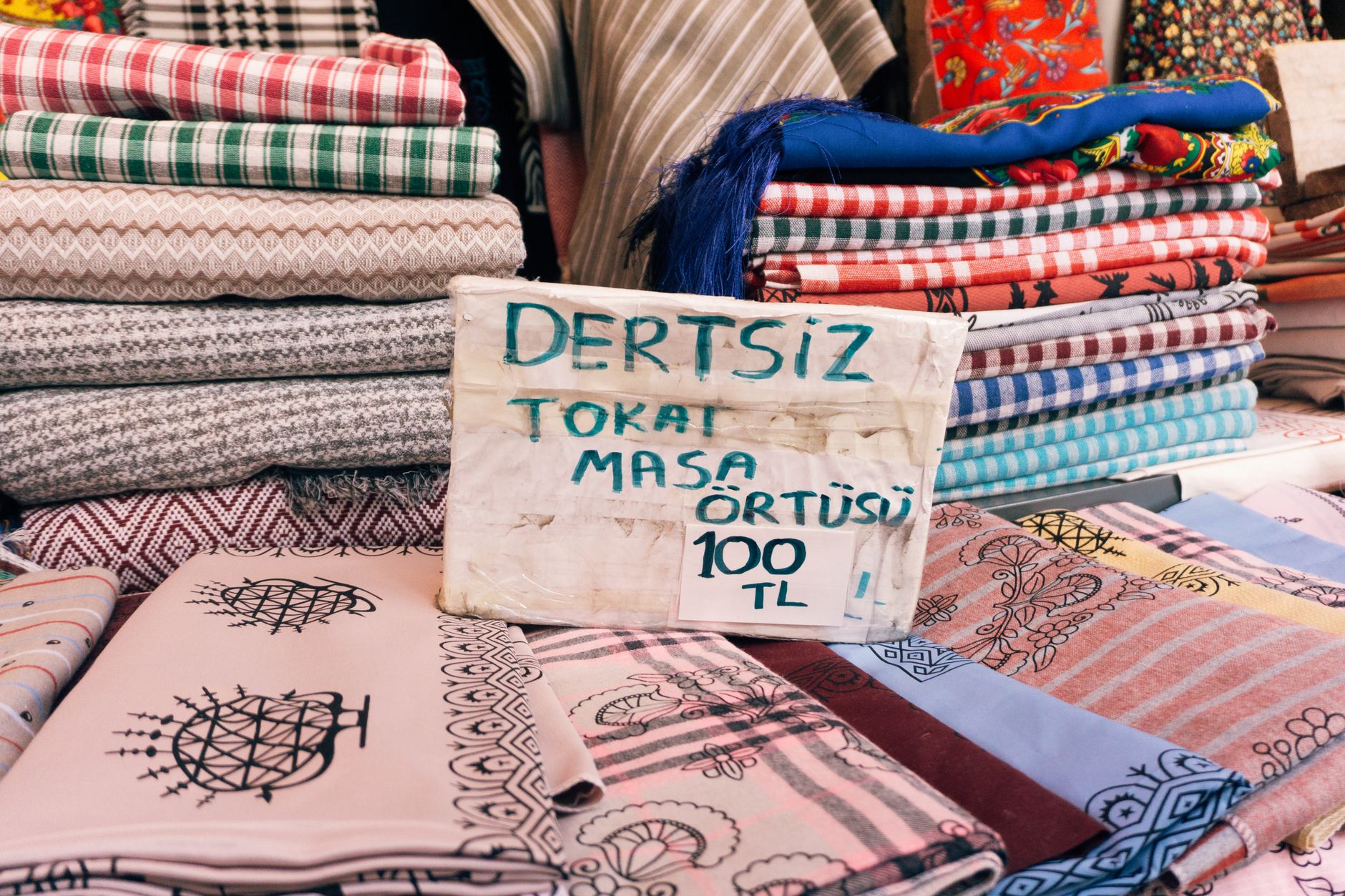 A variety of colorful fabrics and a table cover priced at 100 TL displayed in a bazaar stall.