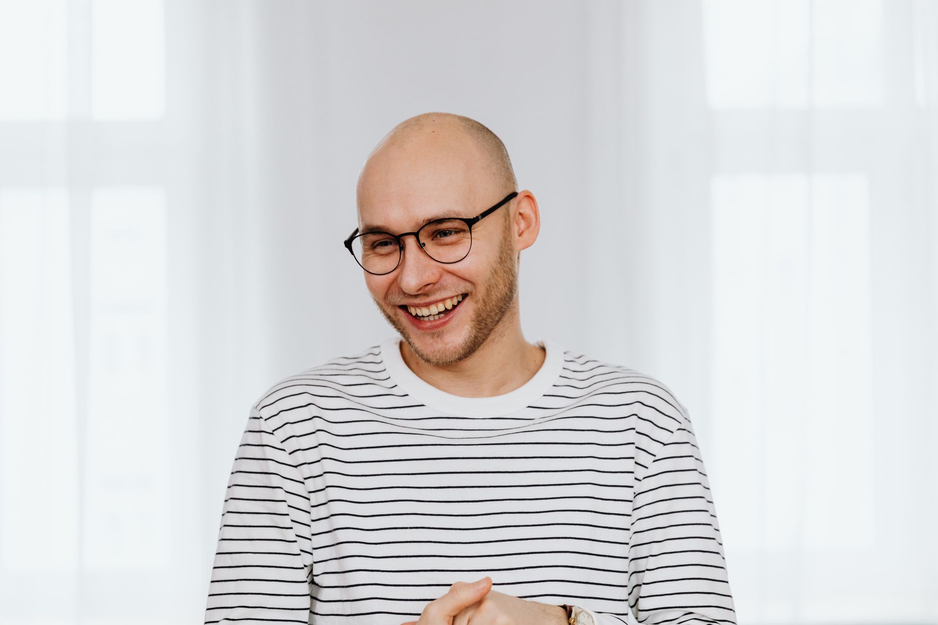 A happy man with a shaved head and glasses, smiling in a bright room.