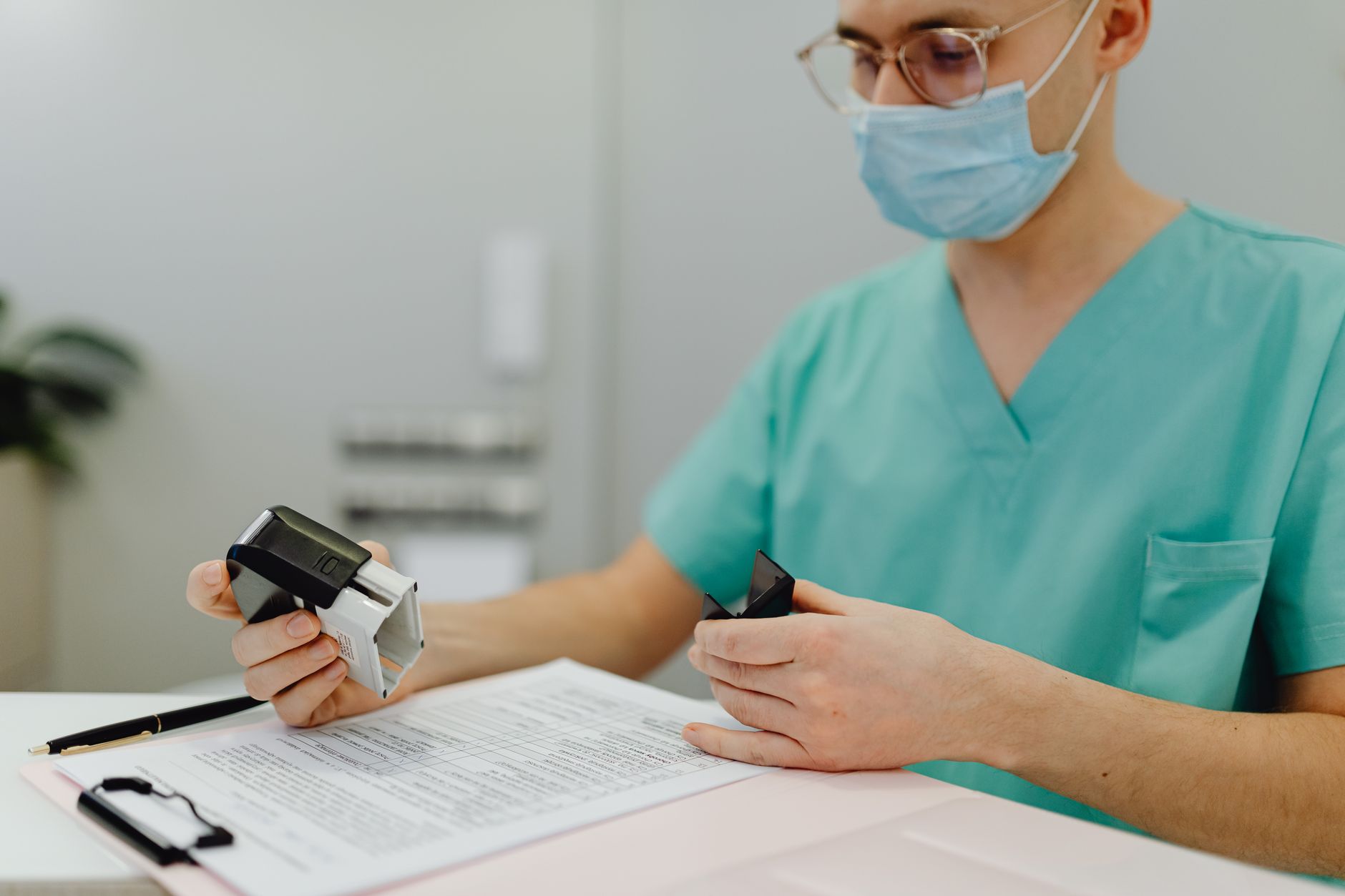 A healthcare worker in scrubs examines medical documents and a device at their desk.