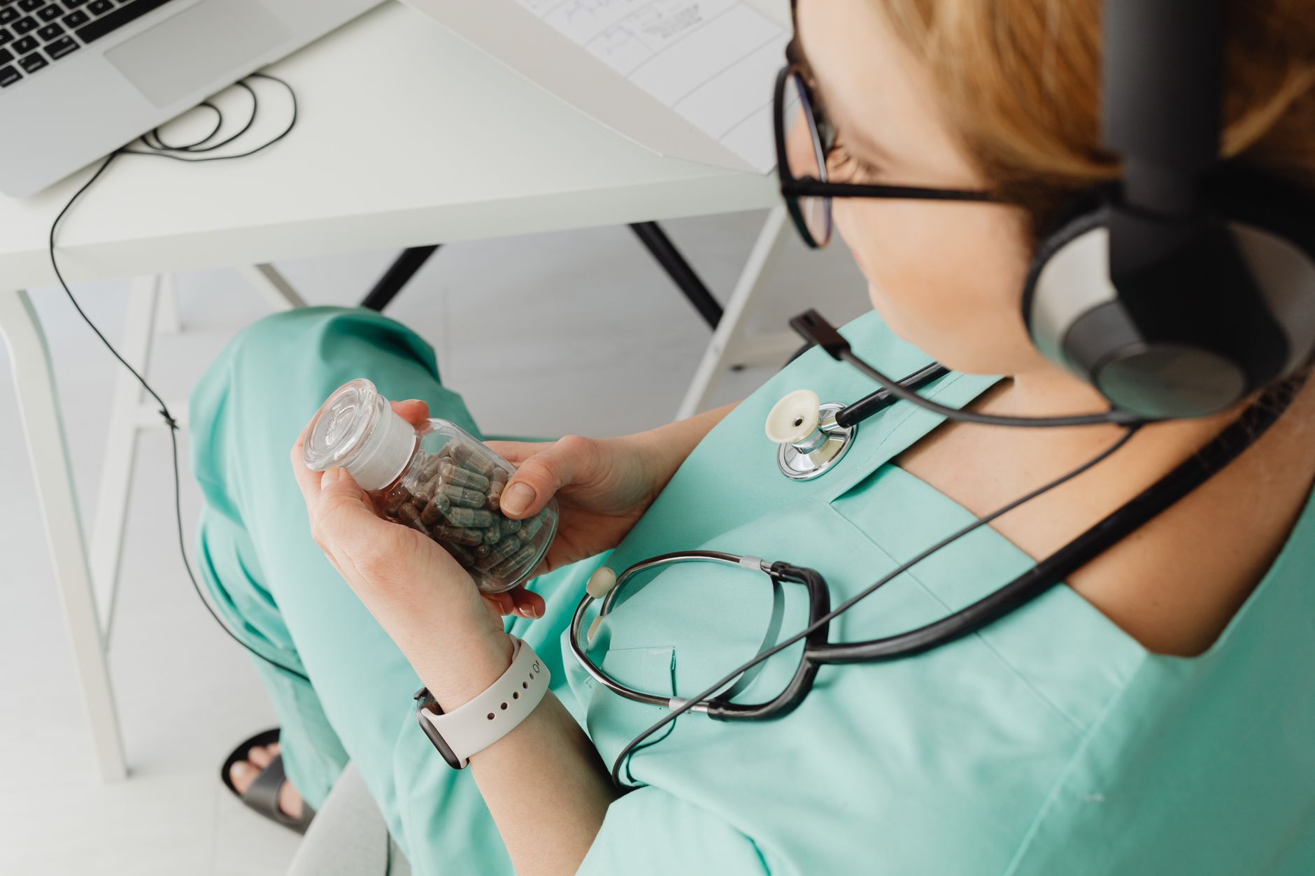 A healthcare worker in teal scrubs holding a bottle of pills while using a headset.