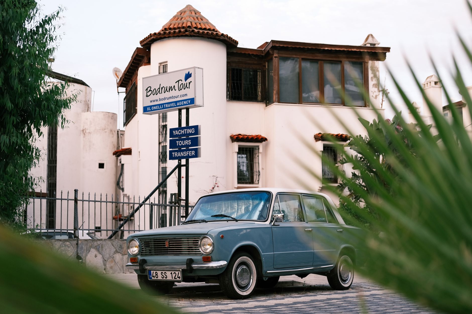 A vintage blue car parked in front of Bodrum Tour agency, featuring yachting and transfer services.
