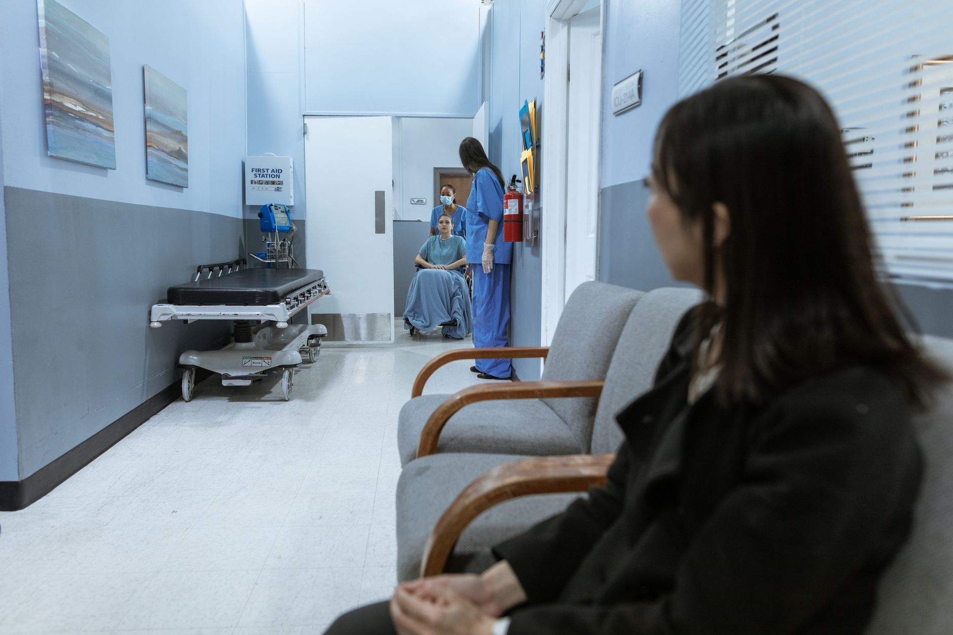 A family member waiting in a hospital lounge while medical staff assist a patient in a wheelchair.