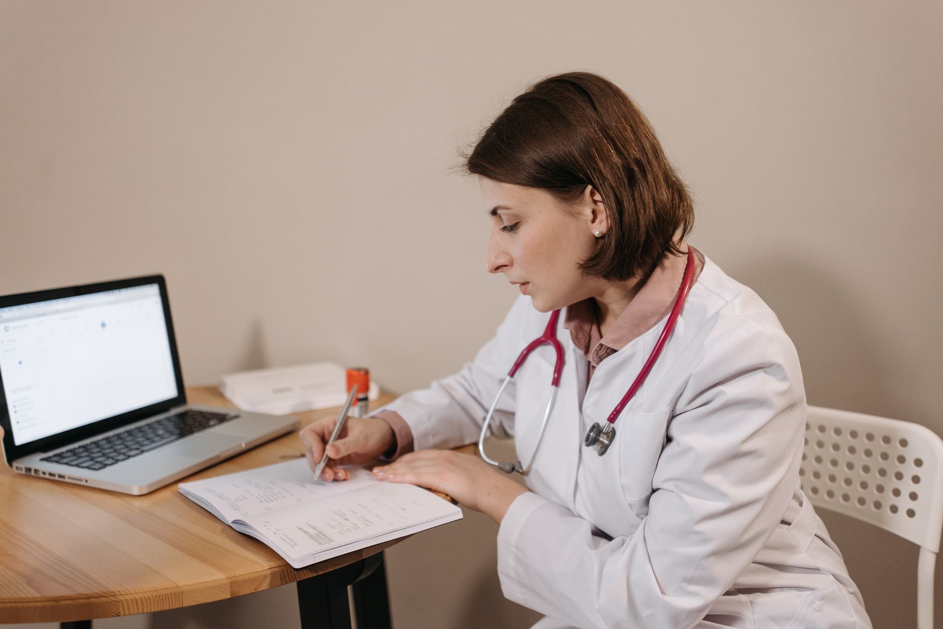 Doctor in white coat taking notes while reviewing digital medical records at a desk.