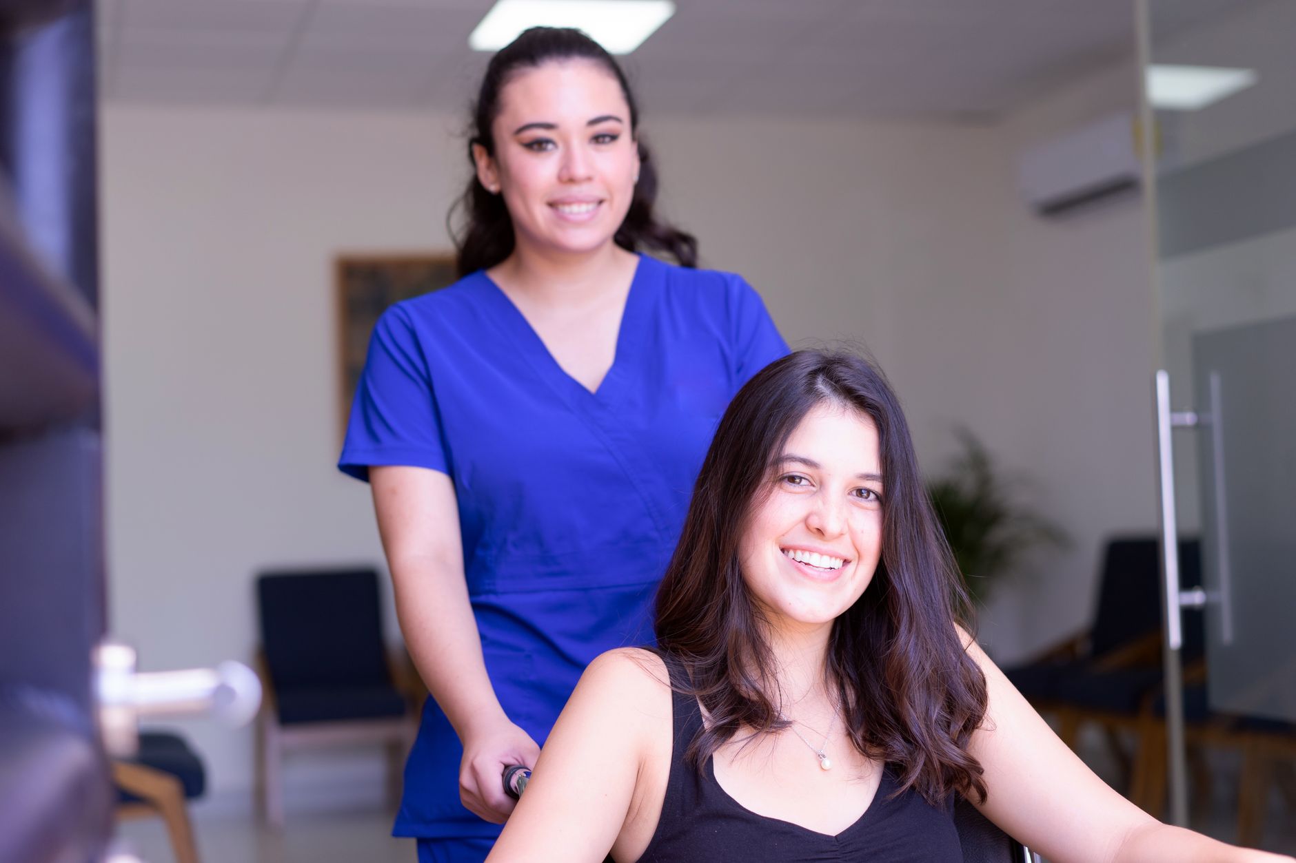 Two women in a hospital setting, one in scrubs pushing a wheelchair with a smiling patient.