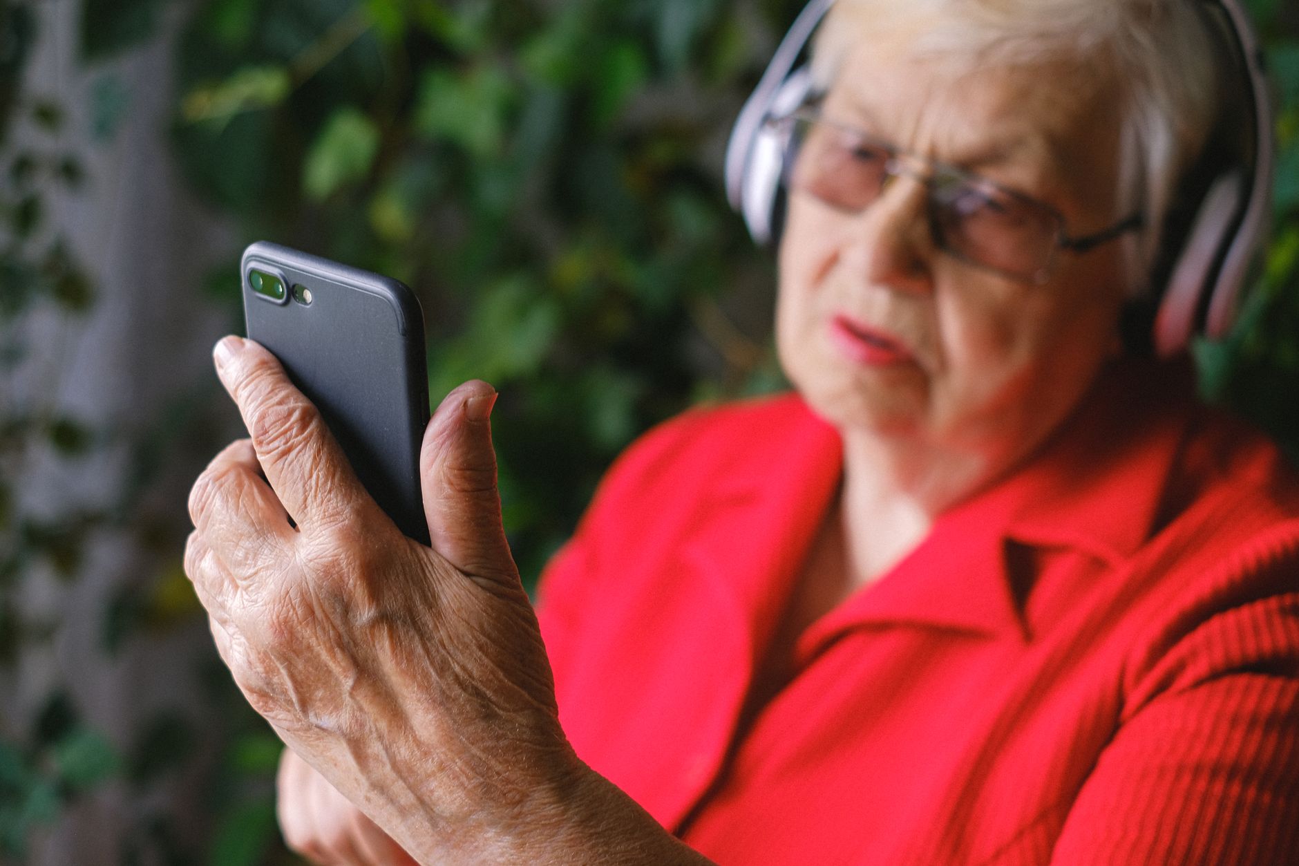 Senior woman in red jacket adjusting hearing aid settings on smartphone.
