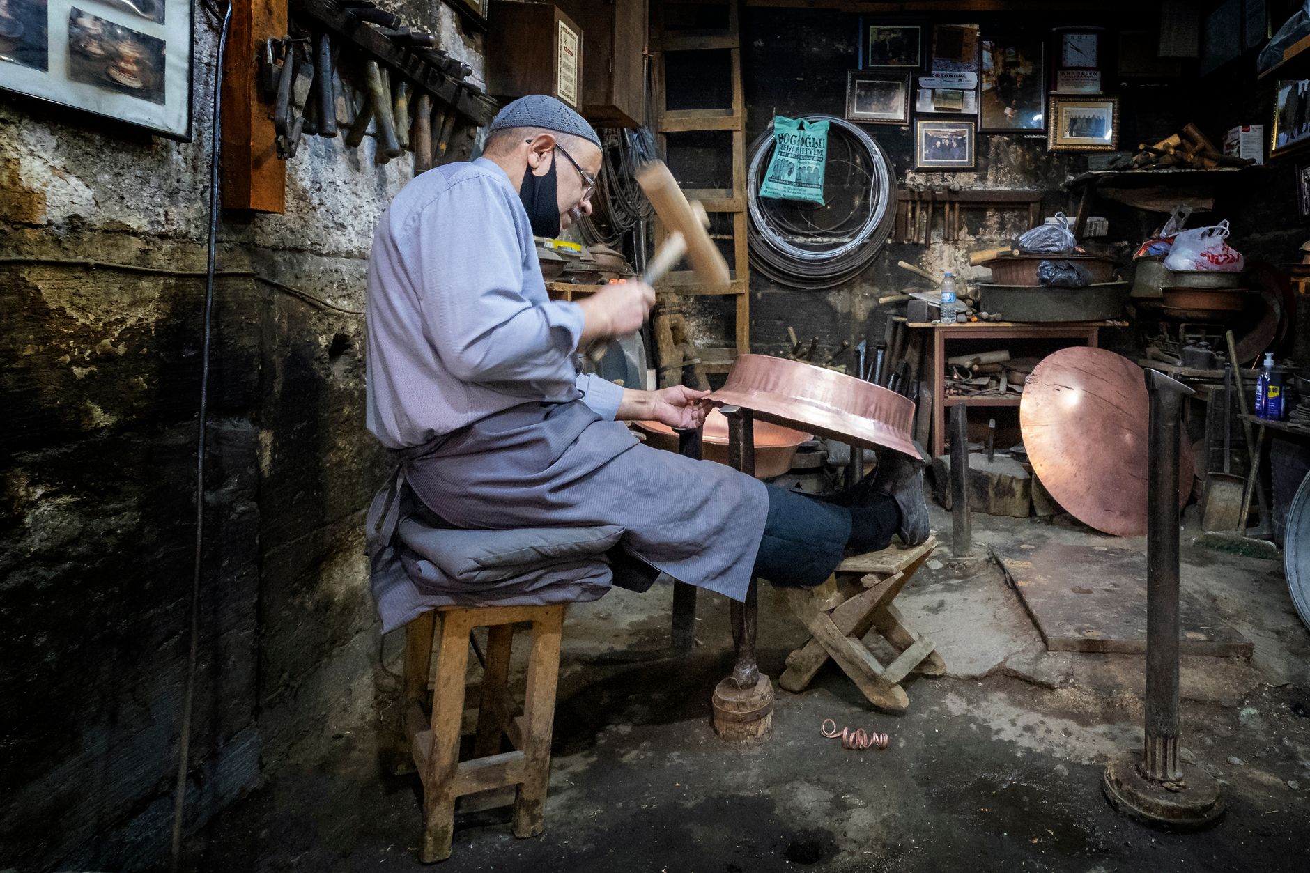 A Turkish artisan skillfully hammers copper in a quaint workshop, surrounded by tools and copper pieces.