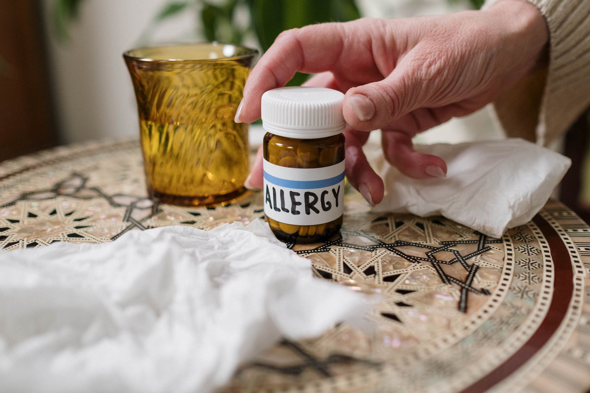 A nurse's hand reaches for a bottle labeled 'ALLERGY' on a decorative table with tissues and a drink.