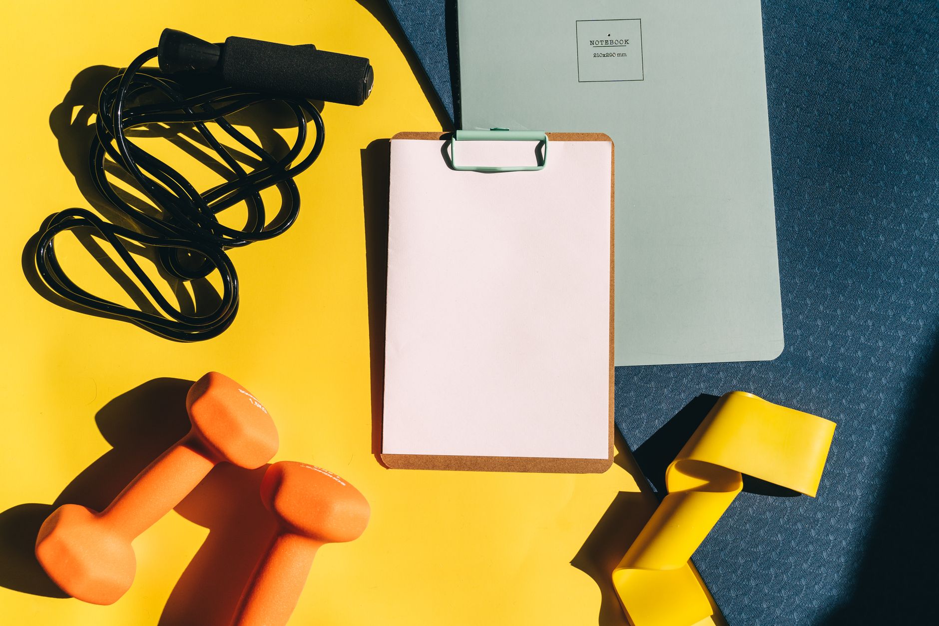 Dumbbells, a jump rope, and a notepad on a yellow surface for home exercise planning.