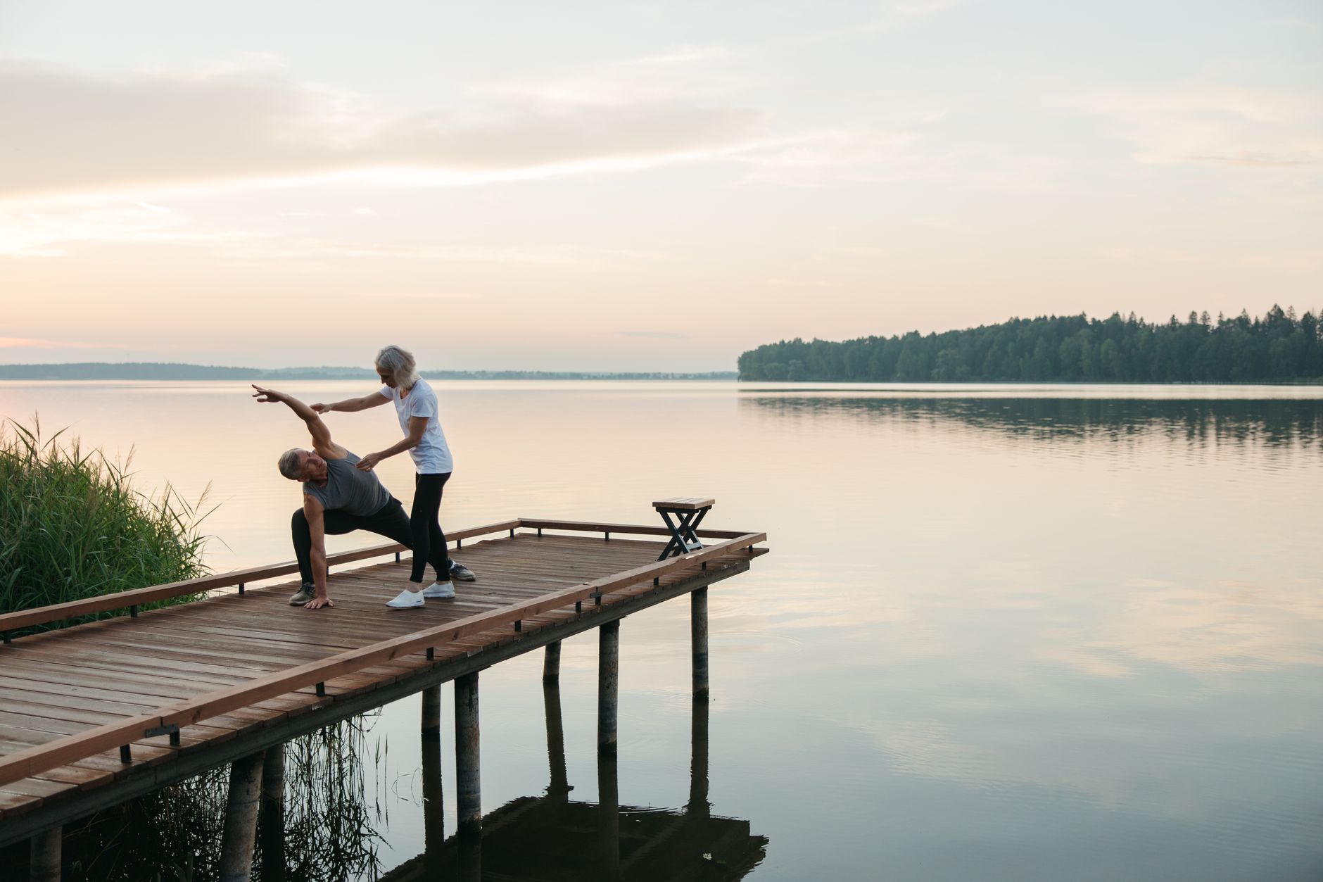 A physical therapist assists a senior man with exercises on a wooden pier by a tranquil lake.